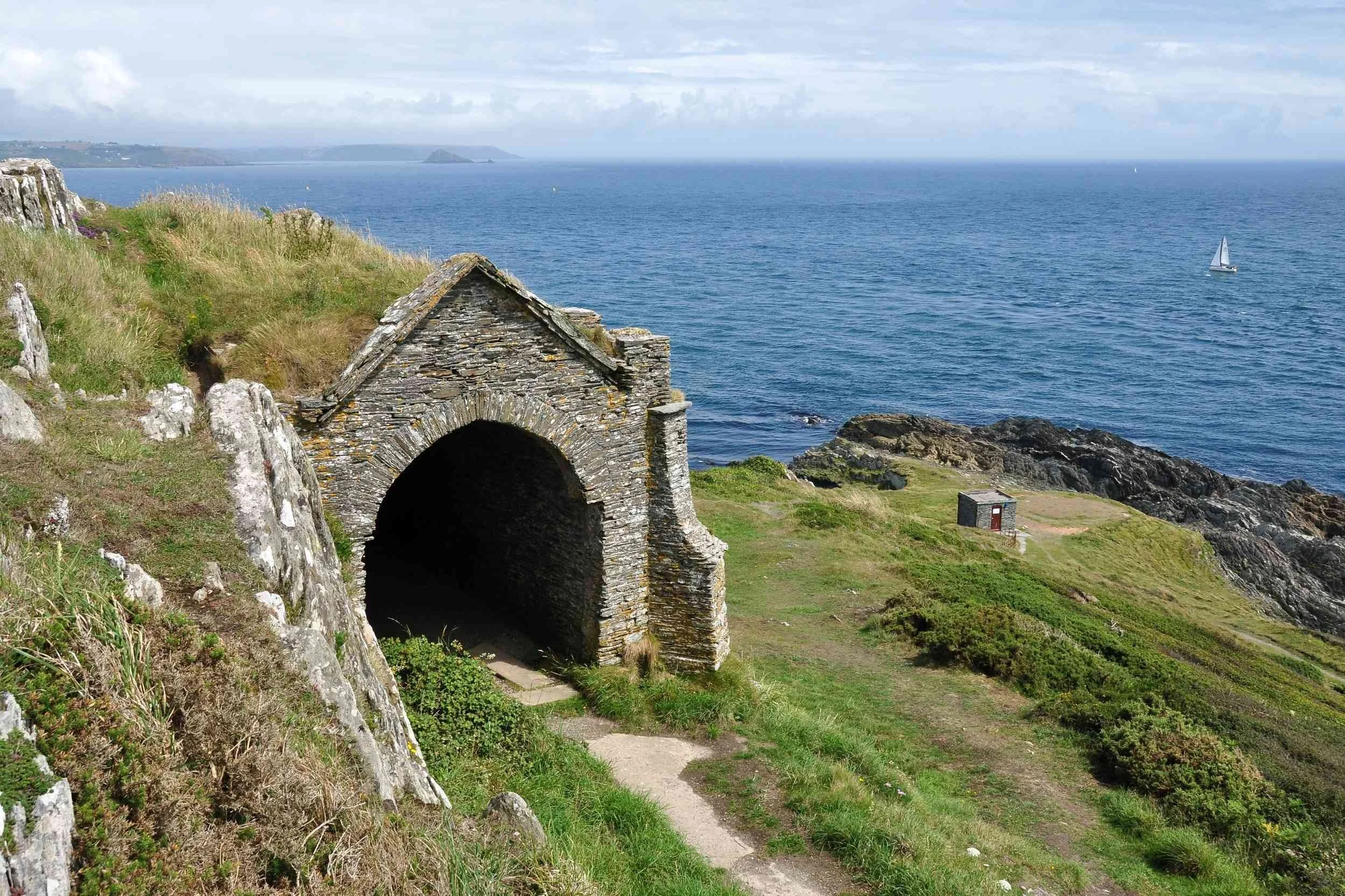 Colour photo of Queen Adelaide’s Grotto at Penlee Point. The lichen covered stonework of the grotto is seen against green grass and a backdrop of sparkling blue sea. Everything is bathed in sunshine.