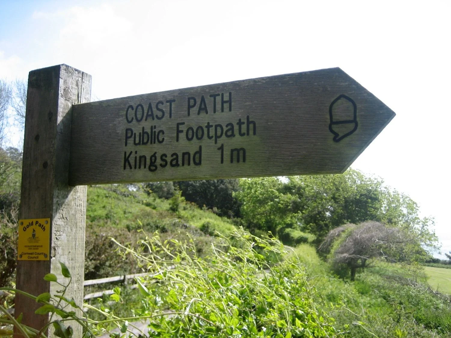 Coast Path signpost with the words 'Public footpath Kingsand 1m'. Shrubs and grass are visible behind the signpost.