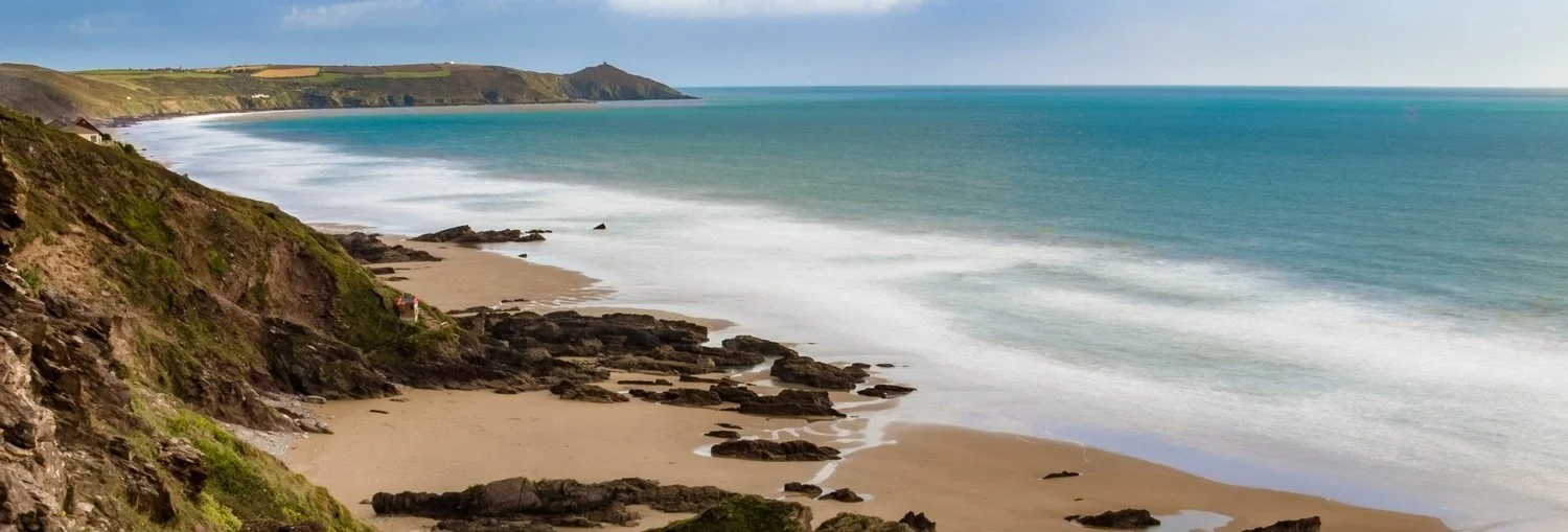 Panoramic view of Whitsand Bay and Rame from west. Sand and rocks in foreground. White surf and blue sea.