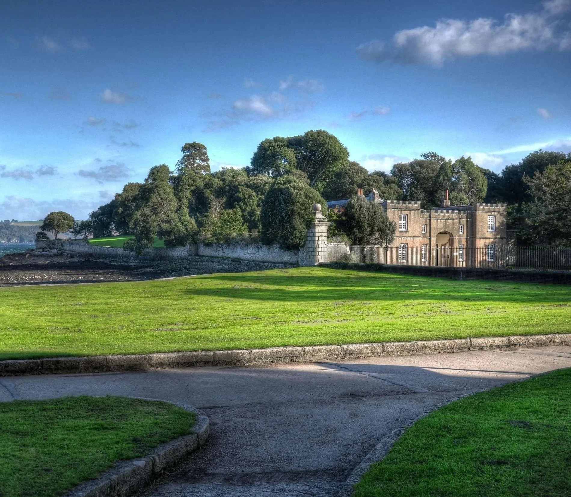 Colour photo of the Country Park boundary viewed from Cremyll.  A castellated former gatehouse, trees and blue sky. Grass in foreground.