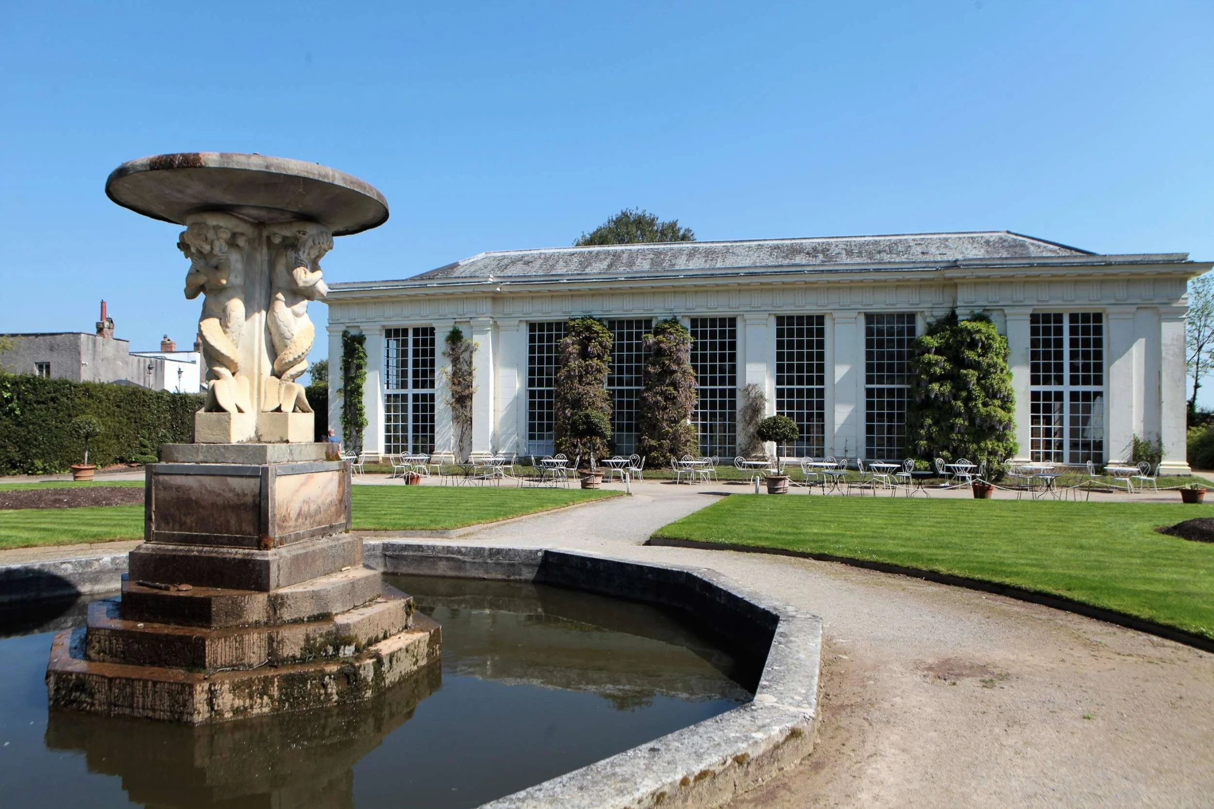 Colour photo of orangery with ornamental pond and fountain in foreground. Blue sky.