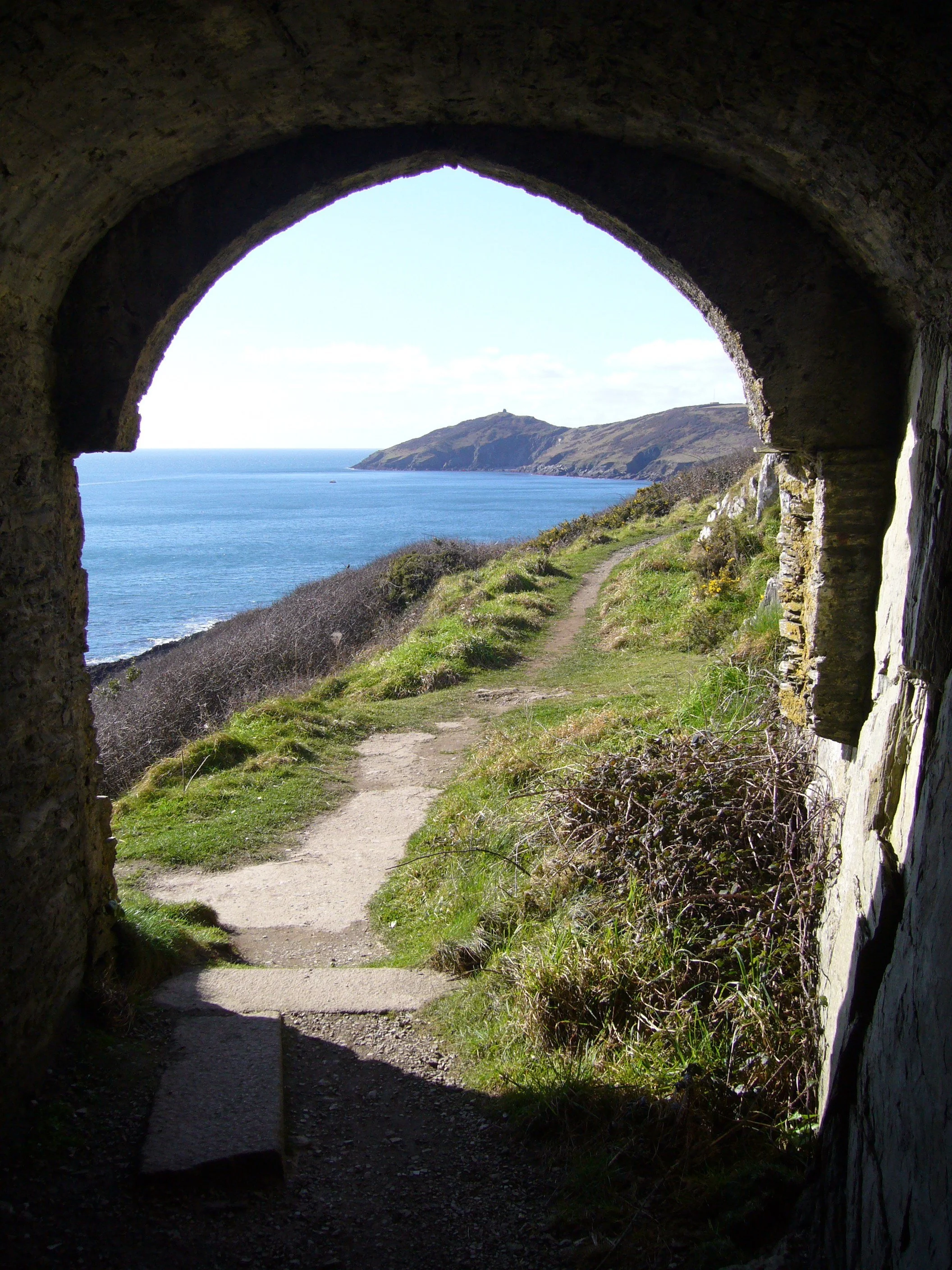 View of headland stretching away to Rame Head. Photo is taken from inside Queen Adelaide's Grotto and framed by an archway. The sea is sparkling blue in the sunshine.