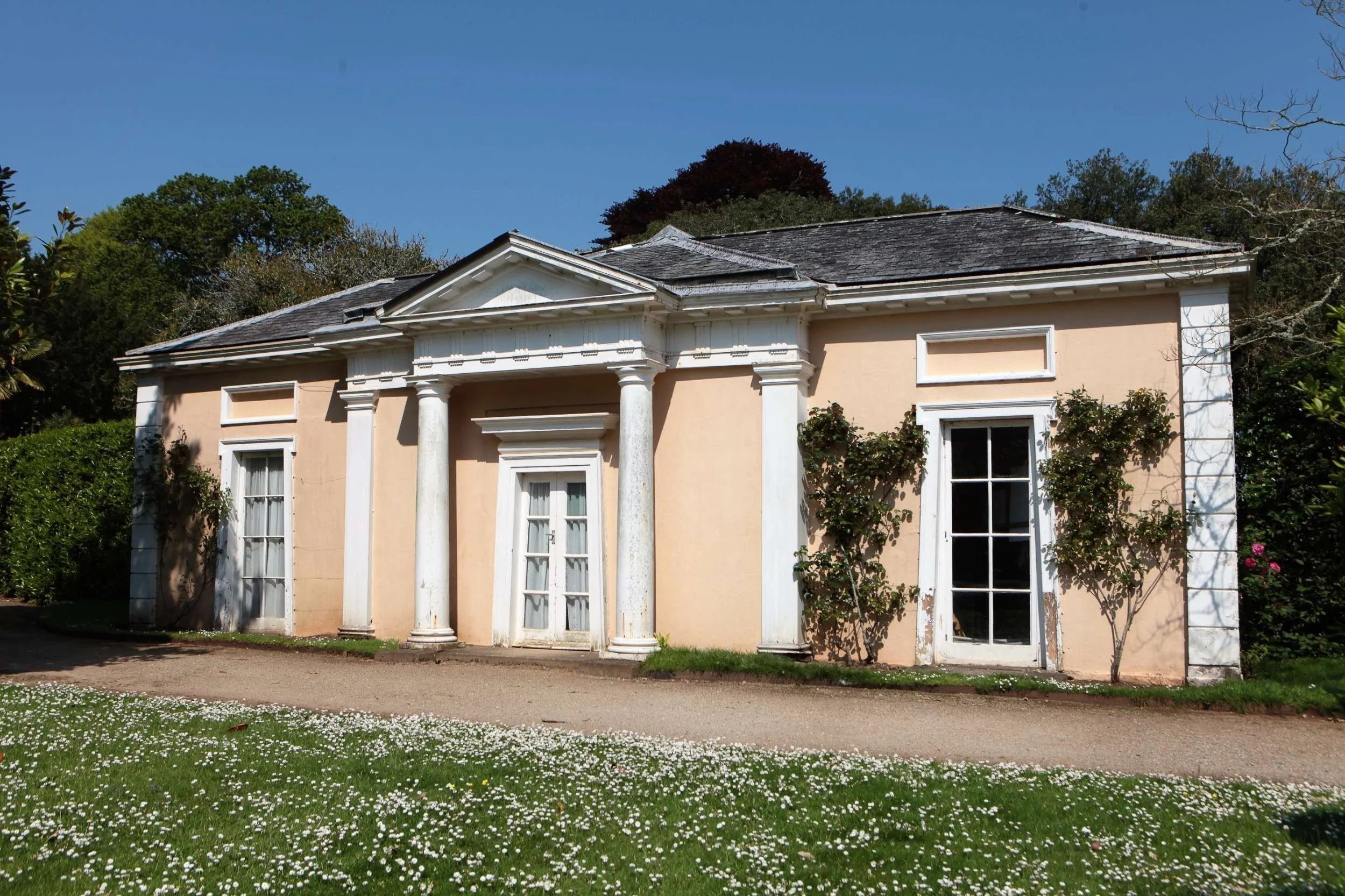 Colour photo of English Garden House with apricot coloured walls and white columns and windows. Blue sky.
