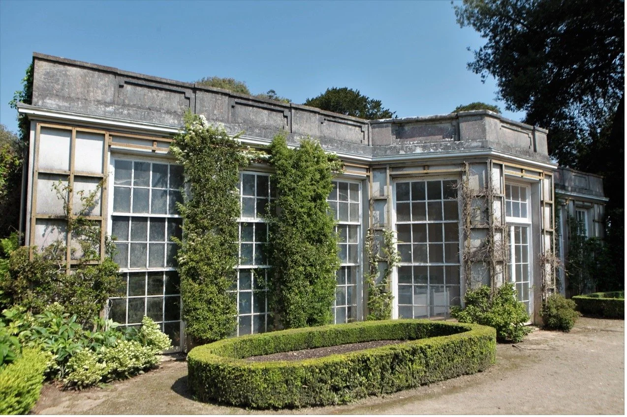 Photo of French Garden House seen from front at an angle.  Between large symmetrical windows trellis supports climbers. Oval box parterre to front of image. Sun and blue sky.