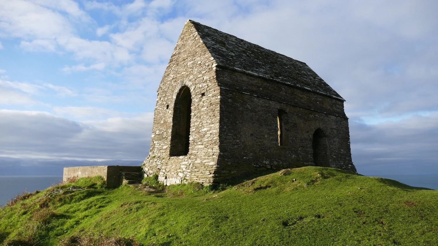 Colour photo of the medieval stone-built Rame Chapel from a few meters away on a sunny day. Close cropped grass is in the foreground and the backdrop is mainly blue sky with fluffy white clouds.