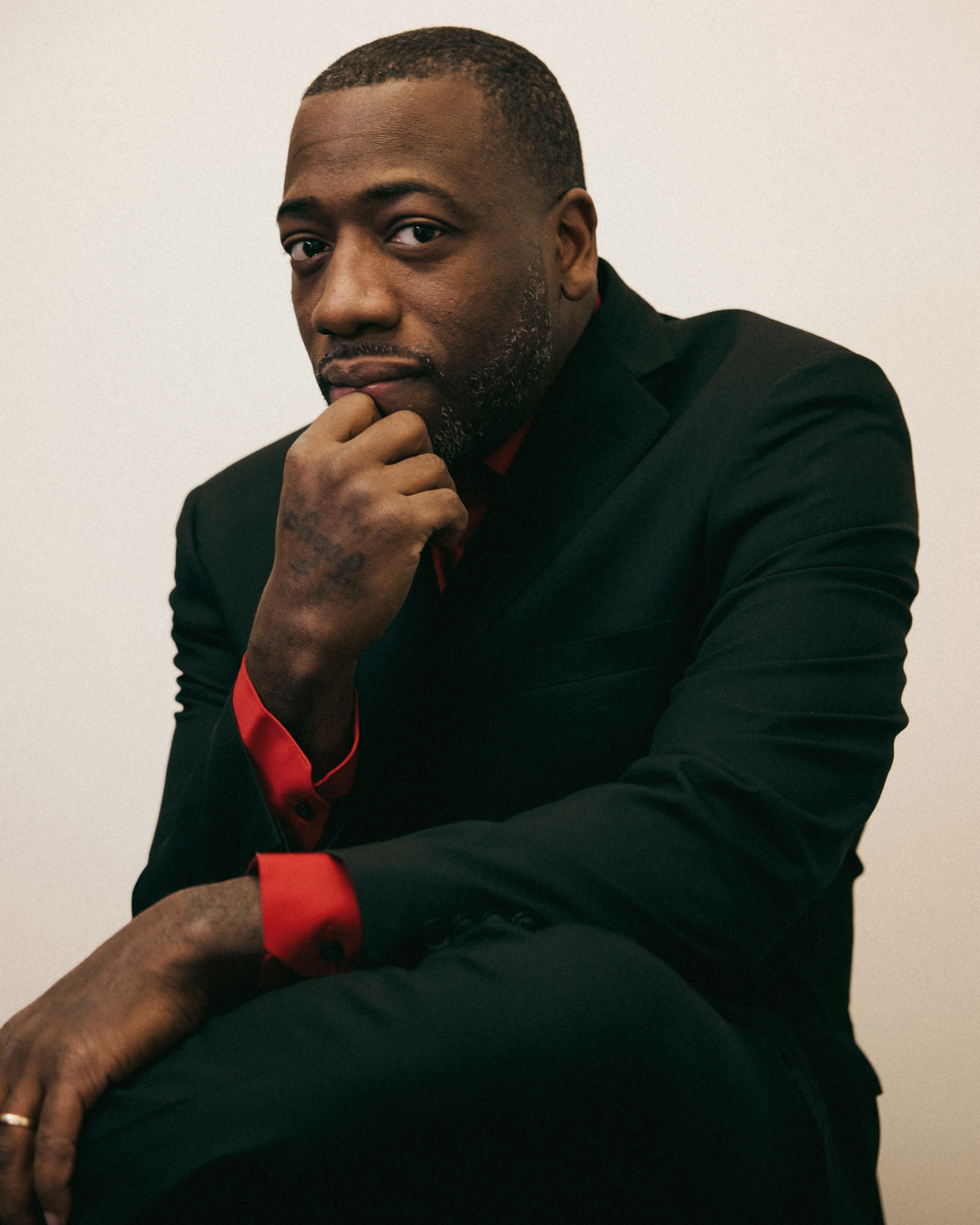 A man with short hair and a beard, wearing a black suit with red shirt accents, sitting against a plain wall, with his hand resting on his chin and looking at the camera.