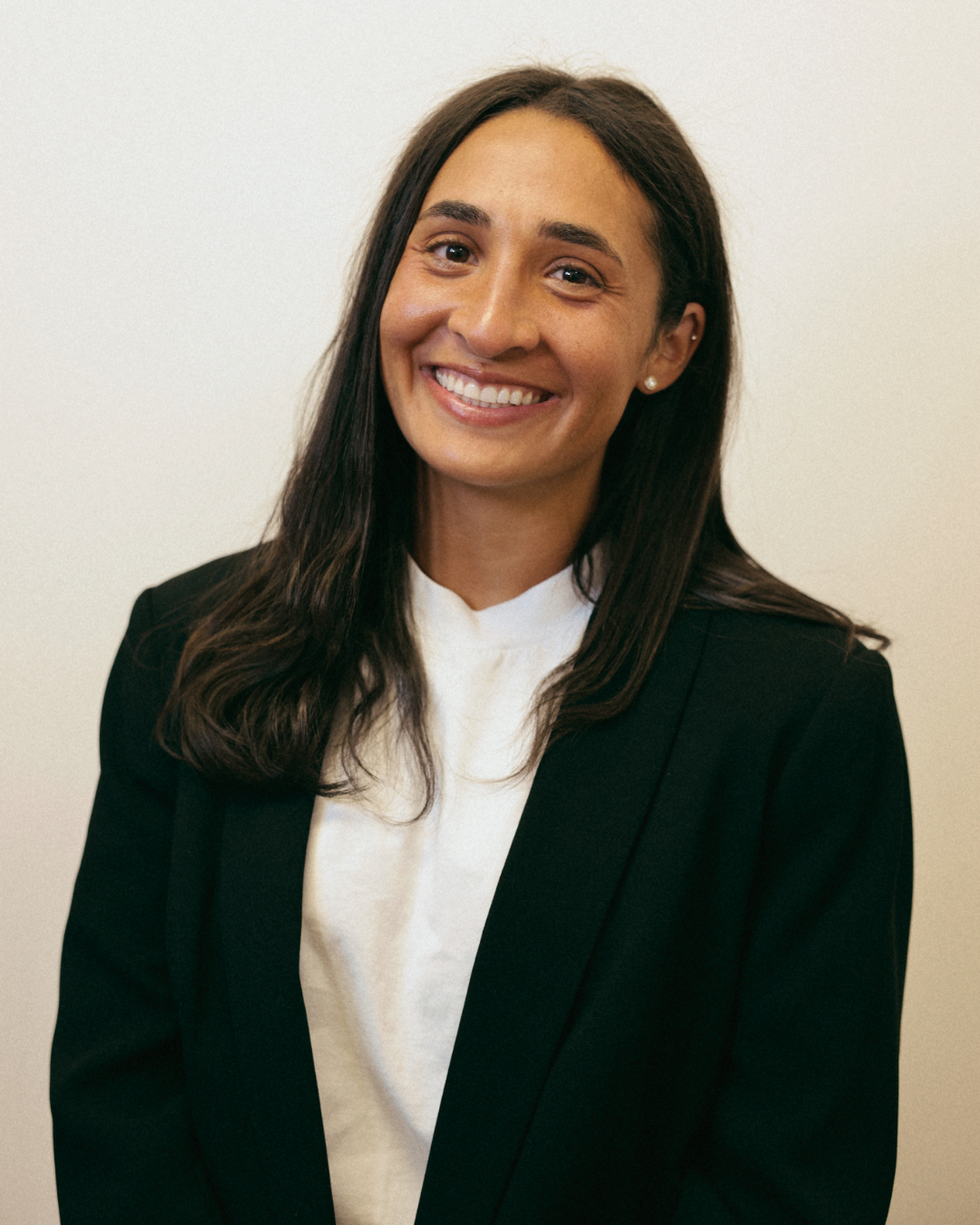 A smiling woman with long dark hair, wearing a black blazer over a white top, standing against a plain light-colored background.