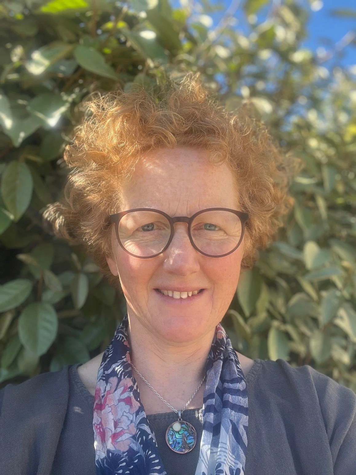A woman with curly red hair, glasses, and a red jacket smiling while taking a selfie in a forested area with trees and fallen leaves.