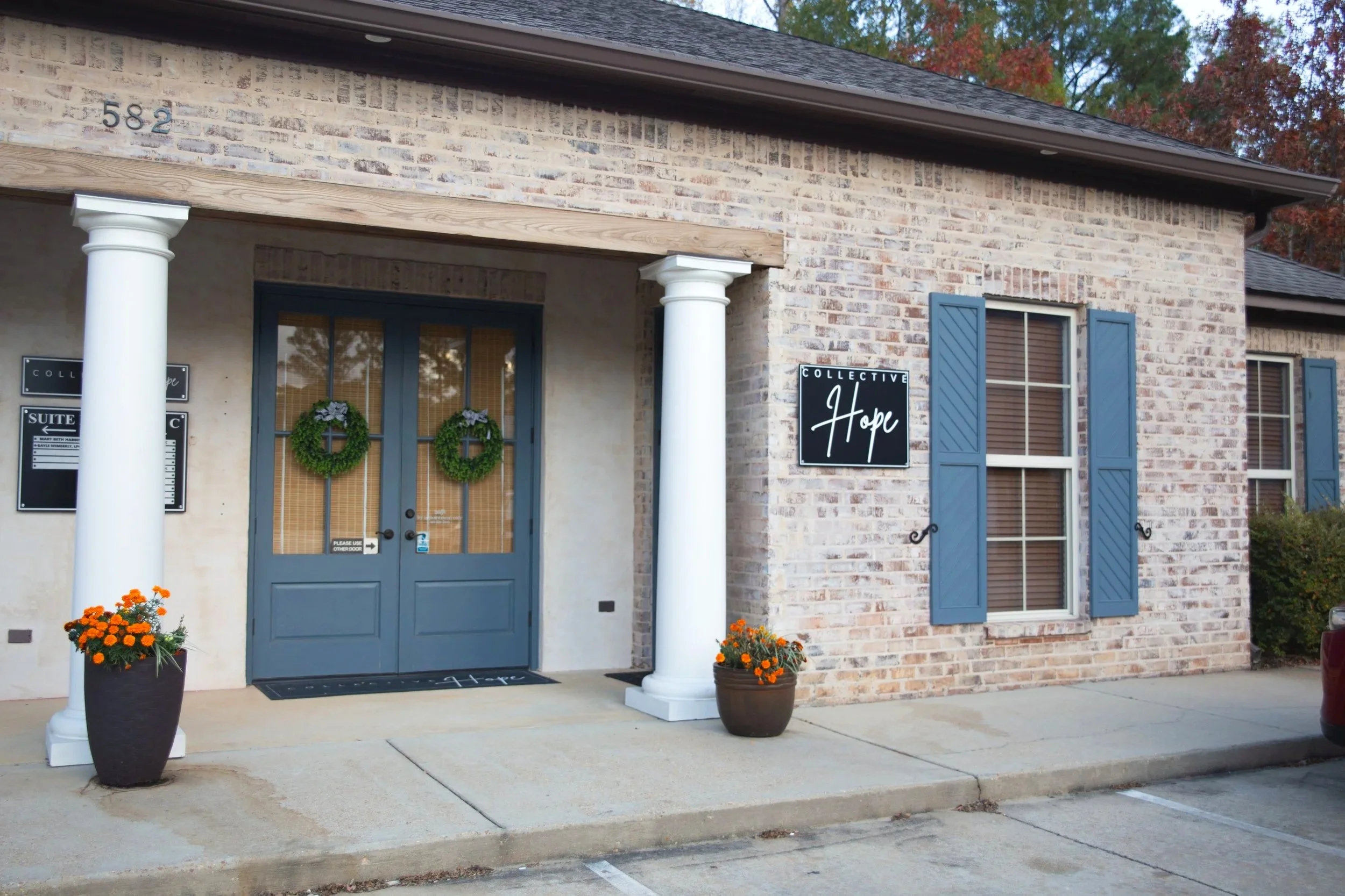 Exterior view of a brick building with blue shutters and a blue door decorated with green wreaths, two potted orange flowers, and signs that read "Collective Hope" and indicating a suite number 582.