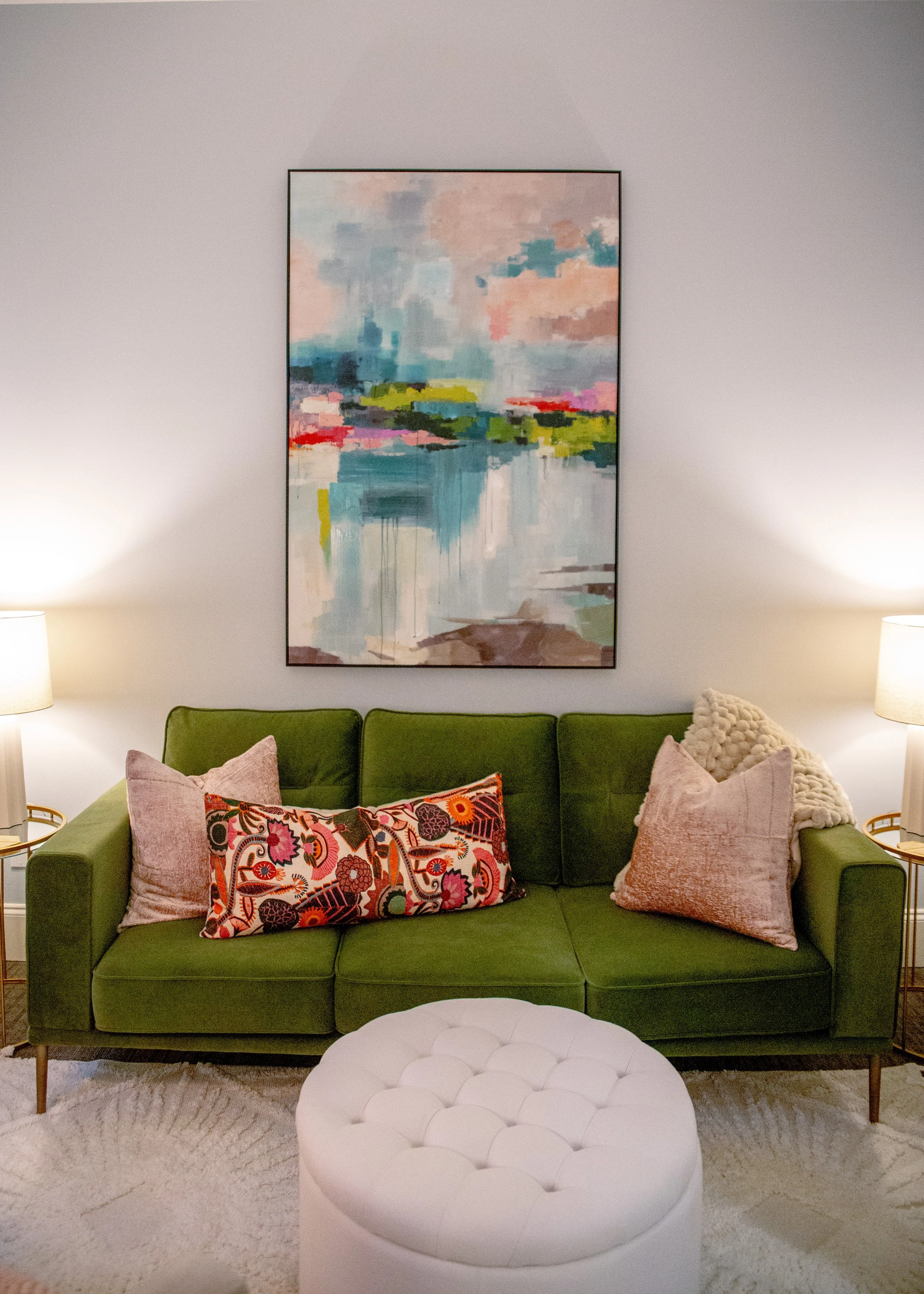 A green sofa with multiple decorative pillows, two lamps on side tables, an abstract painting on the wall, and a white tufted ottoman in front, in a living room.