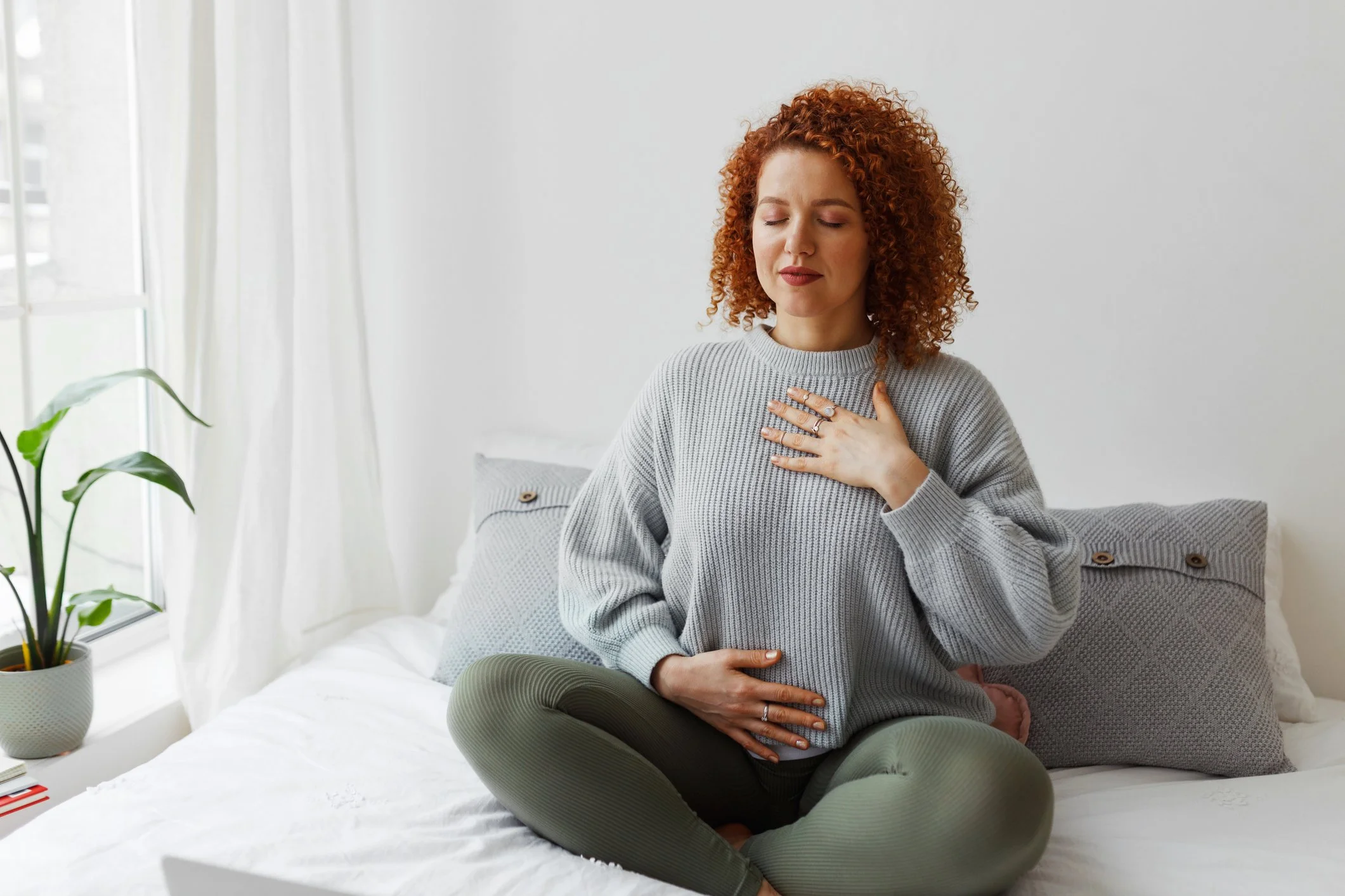 A woman sitting cross-legged on a bed, holding her chest with one hand and her stomach with the other, appearing to be experiencing discomfort or chest pain.