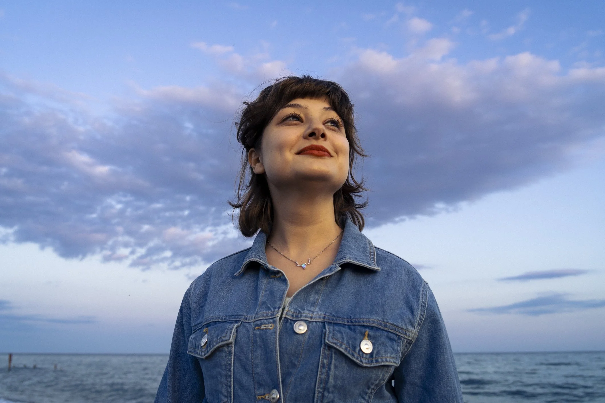 A woman with short brown hair smiling at the ocean with partly cloudy sky in the background, wearing a denim jacket and a necklace.