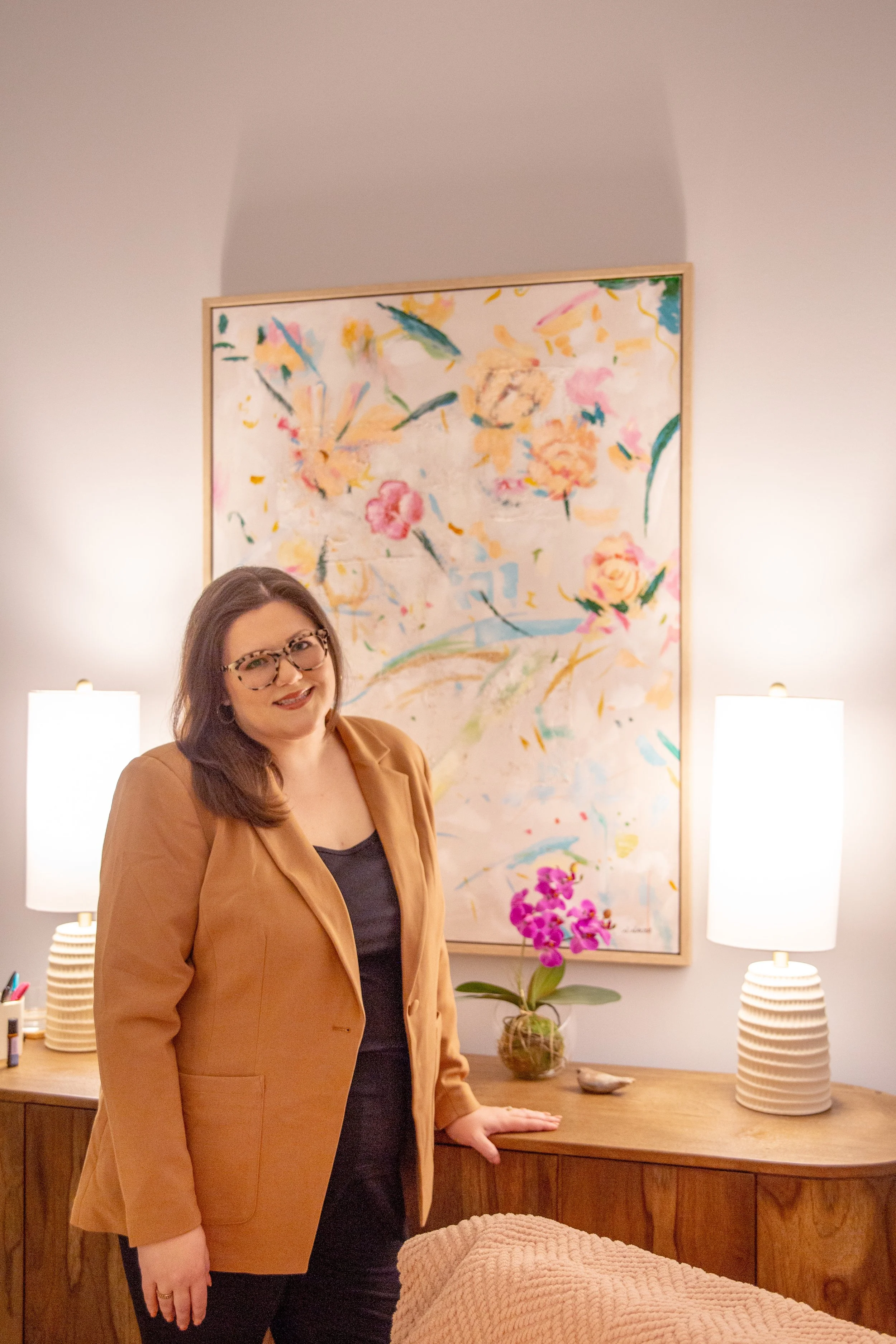 A woman with shoulder-length brown hair, wearing glasses, a black top, and a tan blazer, standing in a softly lit room with two white table lamps, a wooden sideboard, a vase with pink orchids, and a large abstract floral painting on the wall behind her.