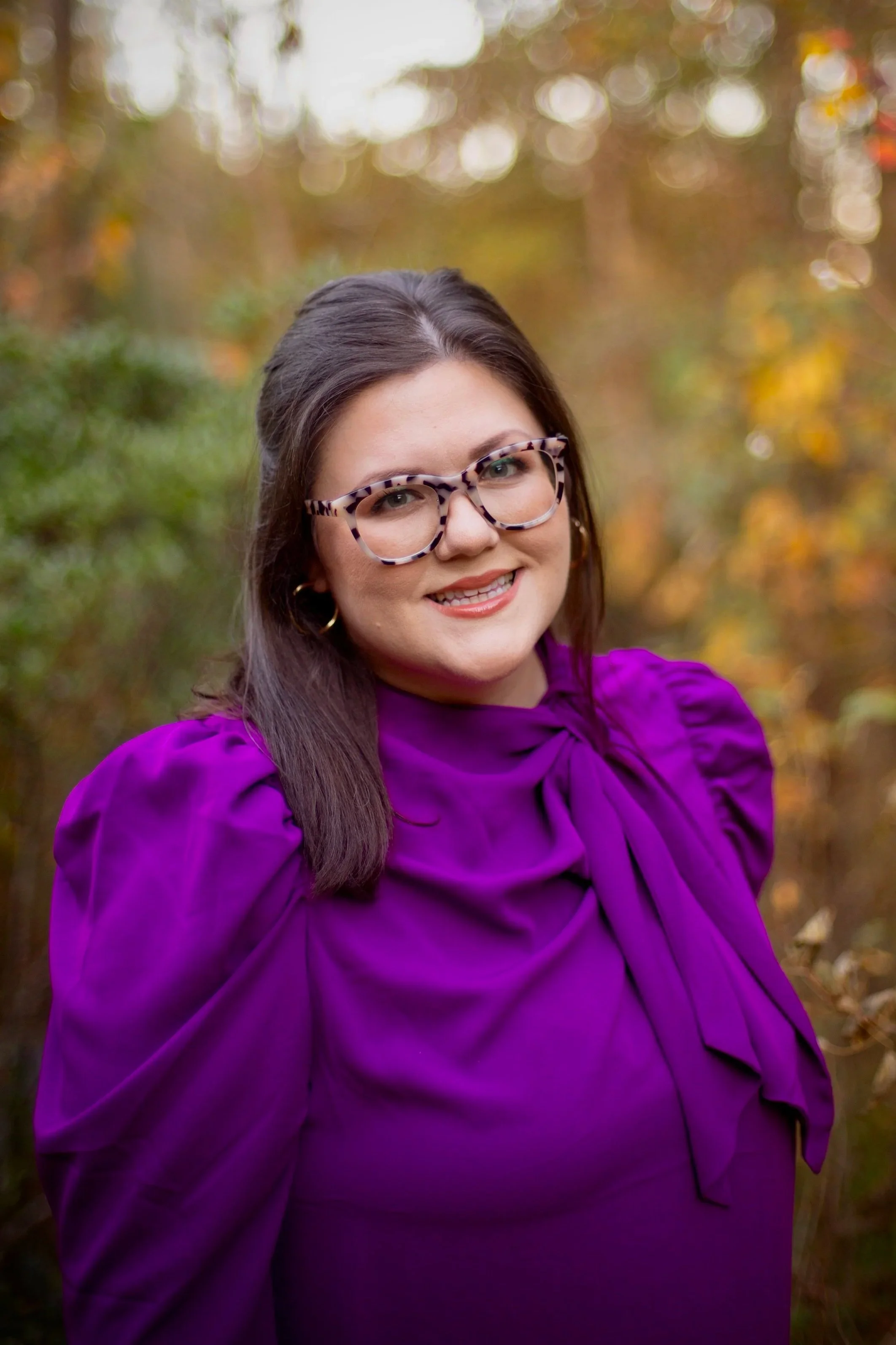 A woman with shoulder-length dark hair, wearing large tortoiseshell glasses and gold hoop earrings, smiling at the camera. She is dressed in a vibrant purple blouse with puffed shoulders and a bow detail at the neck. The background features a blurred natural outdoor setting with trees and autumn-colored leaves.