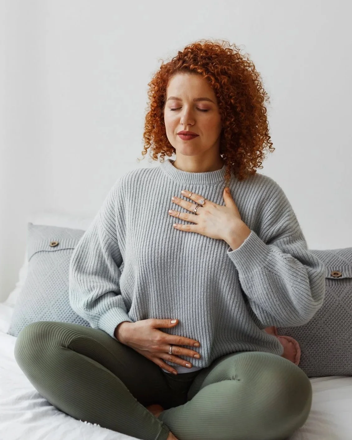A young woman with curly red hair, sitting cross-legged on a bed, wearing a gray sweater and green leggings, with one hand on her chest and the other on her abdomen, appearing to experience a moment of reflection or emotional stress.