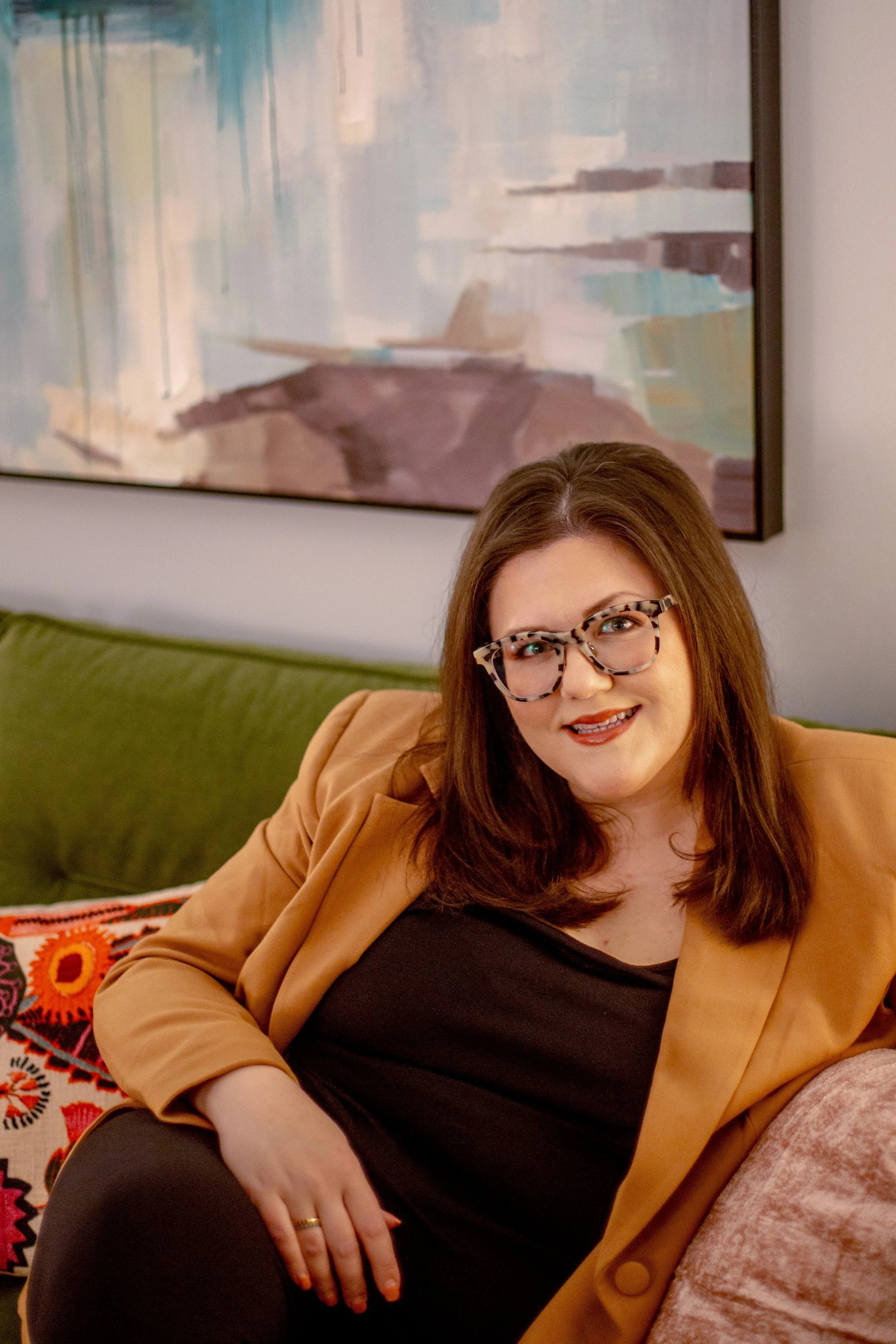A woman with shoulder-length brown hair and glasses, wearing a tan blazer over a black top, sitting on a green couch with a patterned pillow. Behind her is a colorful abstract painting on the wall.