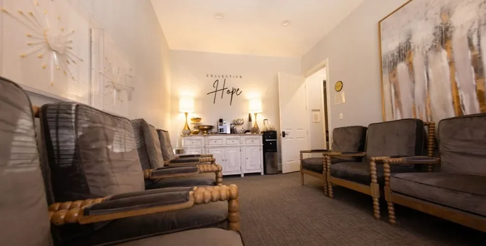 A waiting room with six brown cushioned chairs with wooden armrests and legs, a white sideboard with lamps, bowls, and decor, a large abstract painting on the wall, and a sign that says "Collective Hope".