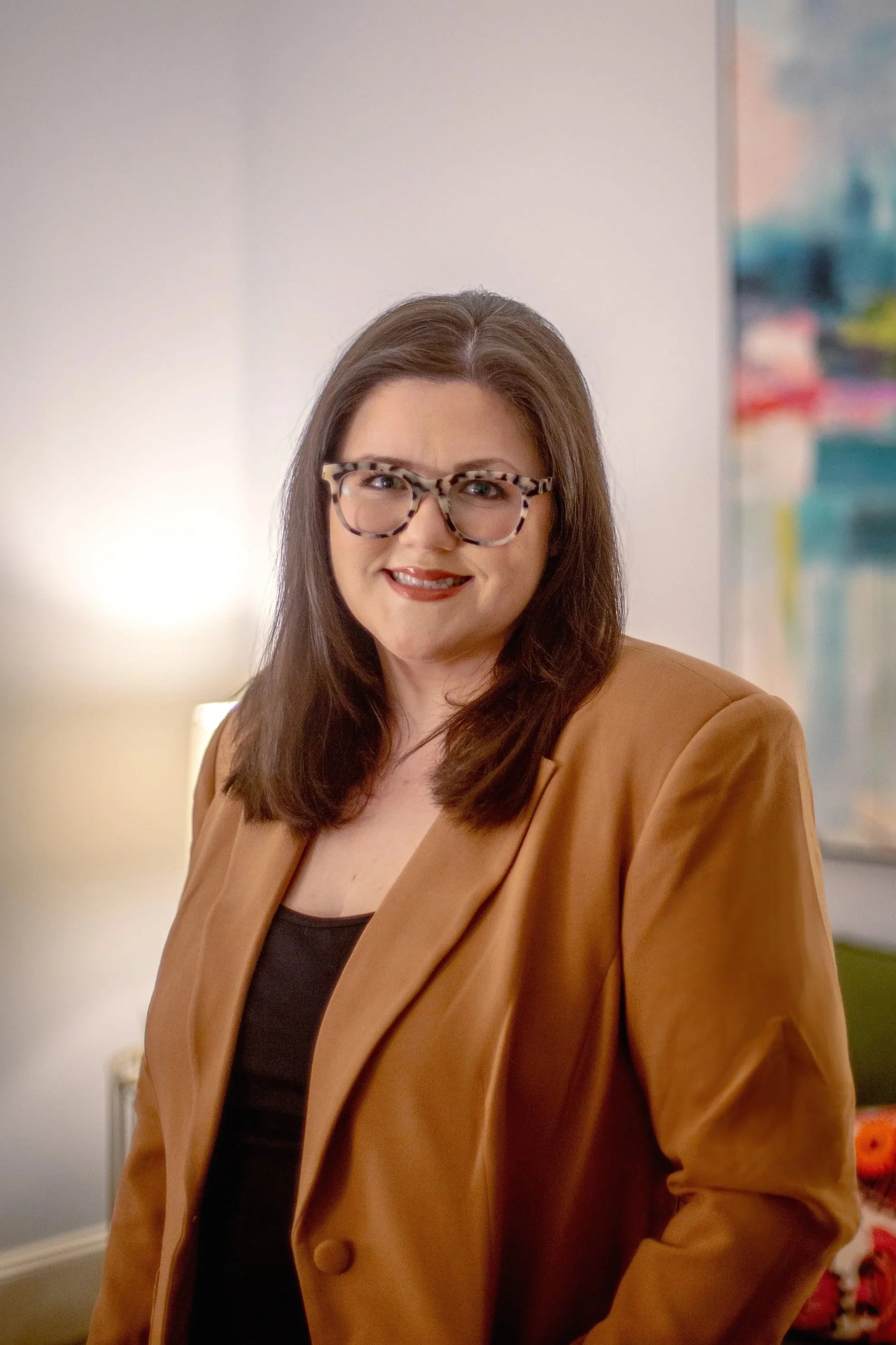 A woman with shoulder-length brown hair, wearing large tortoiseshell glasses, a black top, and a tan blazer, standing indoors with a colorful painting and a lamp in the background.