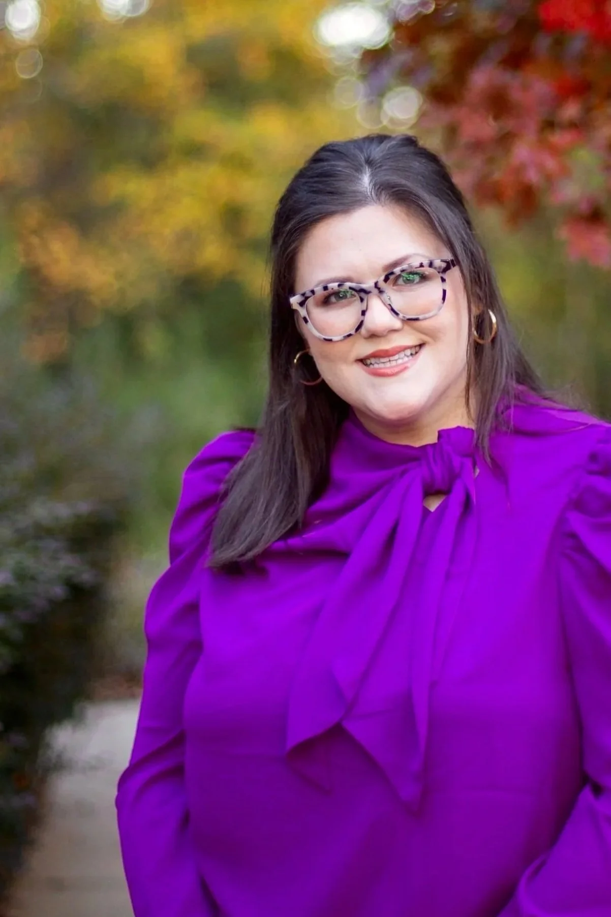 A woman with long dark hair wearing purple blouse with a bow, patterned glasses, and hoop earrings, smiling outdoors with autumn foliage in the background.