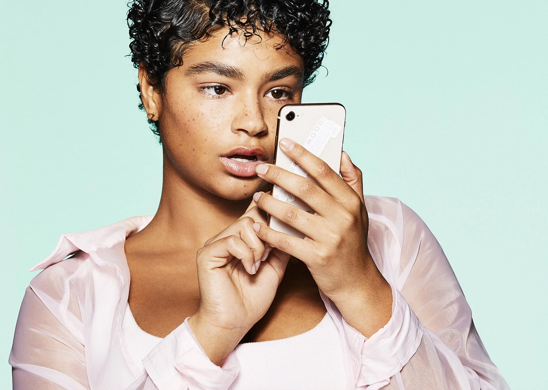 A young woman with curly short hair, wearing a light pink semi-transparent shirt and a white top, looking at her phone with a surprised or curious expression, against a light mint green background.