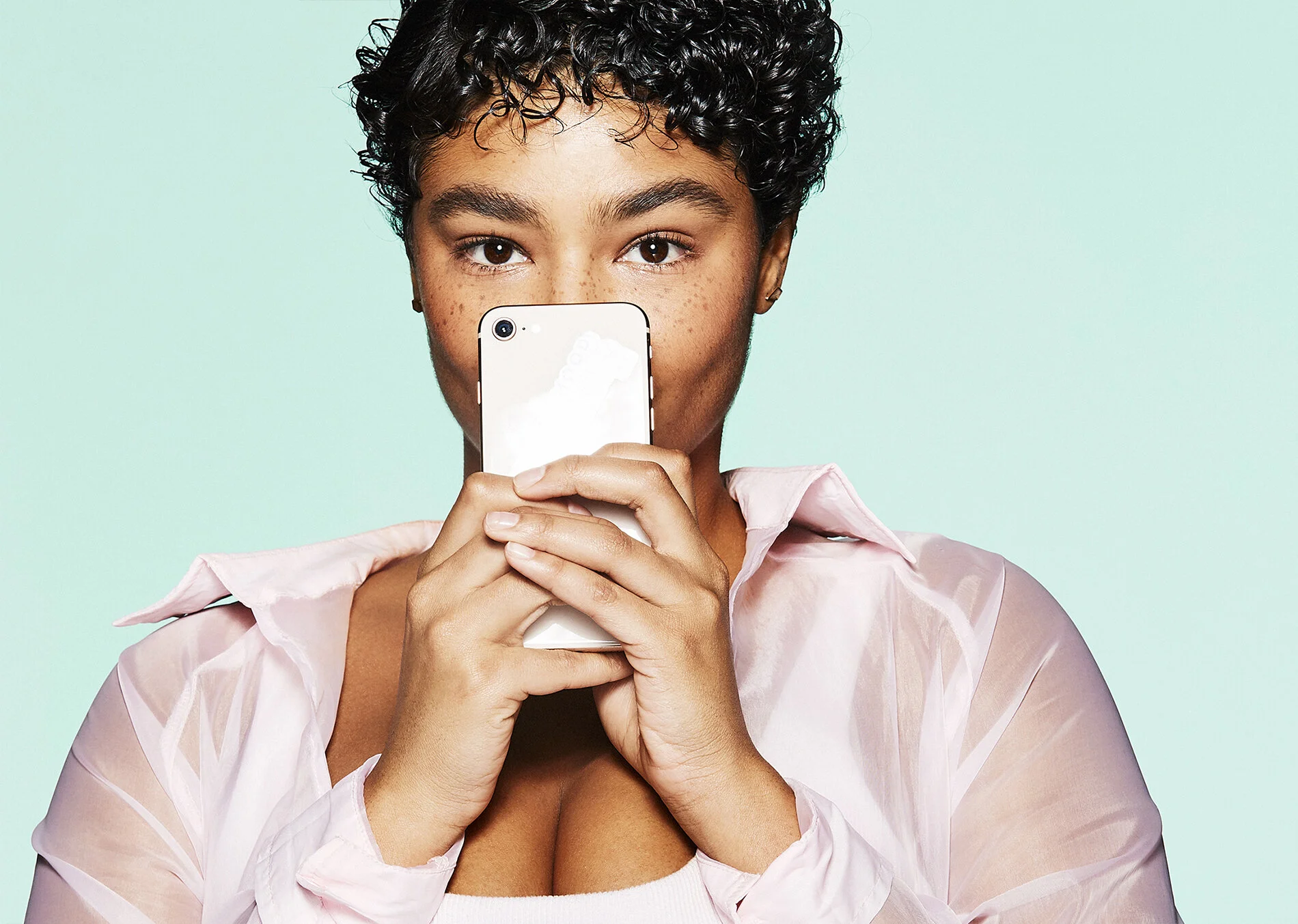 A woman with short, curly hair taking a selfie with her phone, standing against a light pastel background.