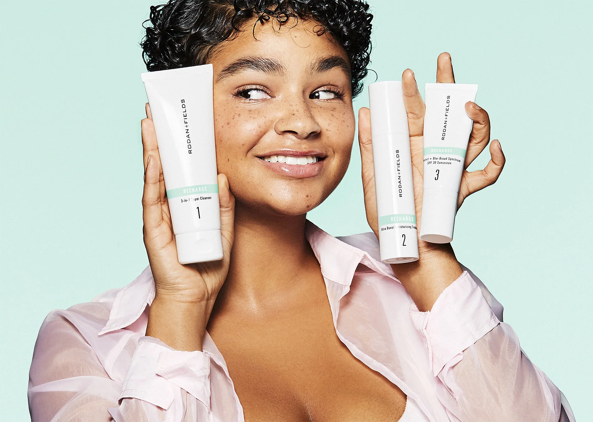 A woman with short, curly hair and freckles smiling while holding three skincare products from Rodan + Fields, labeled for different purposes, against a pastel background.
