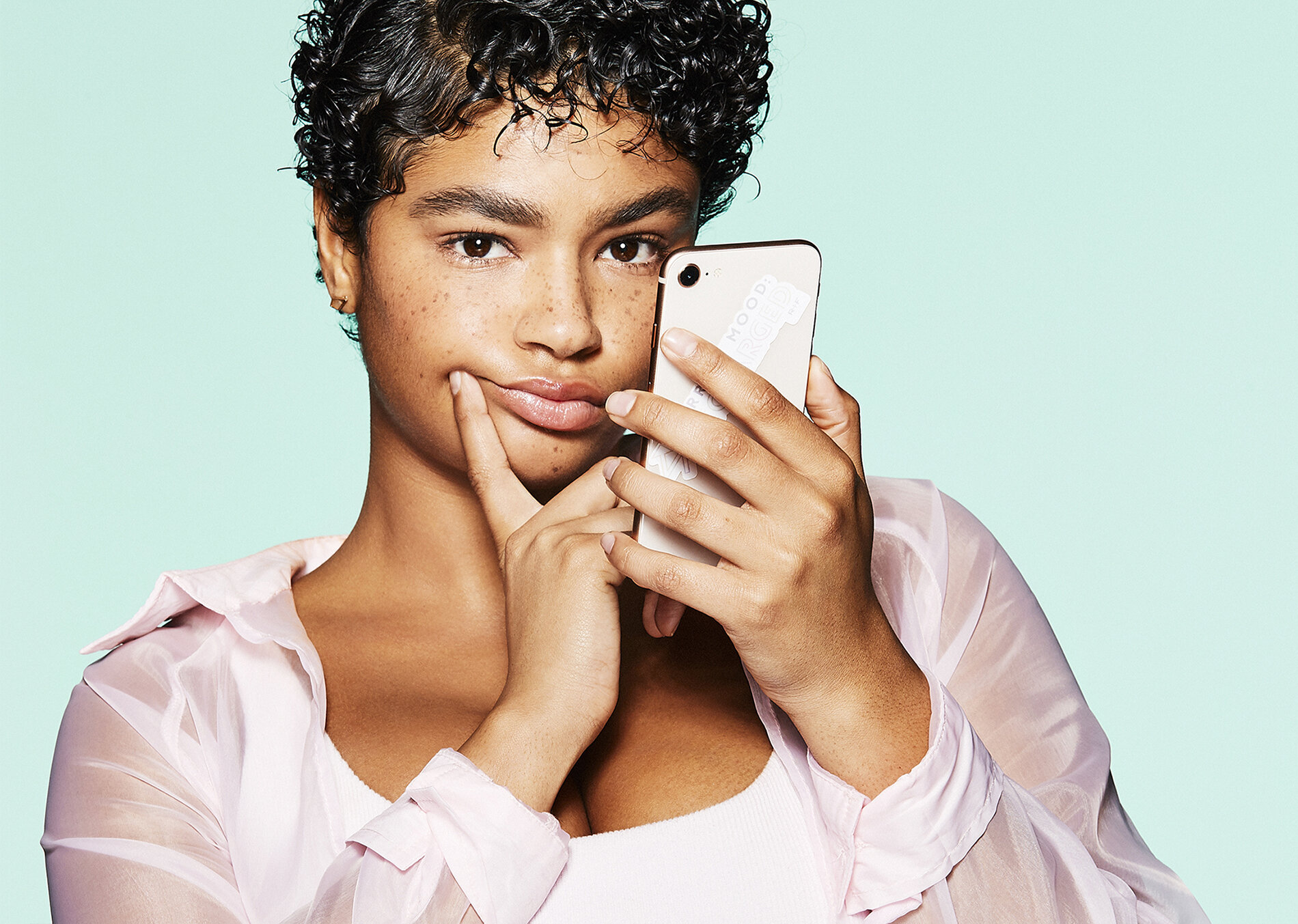 A woman with short, curly hair and a light pink sheer blouse holding a smartphone near her face, touching her chin, against a light green background.