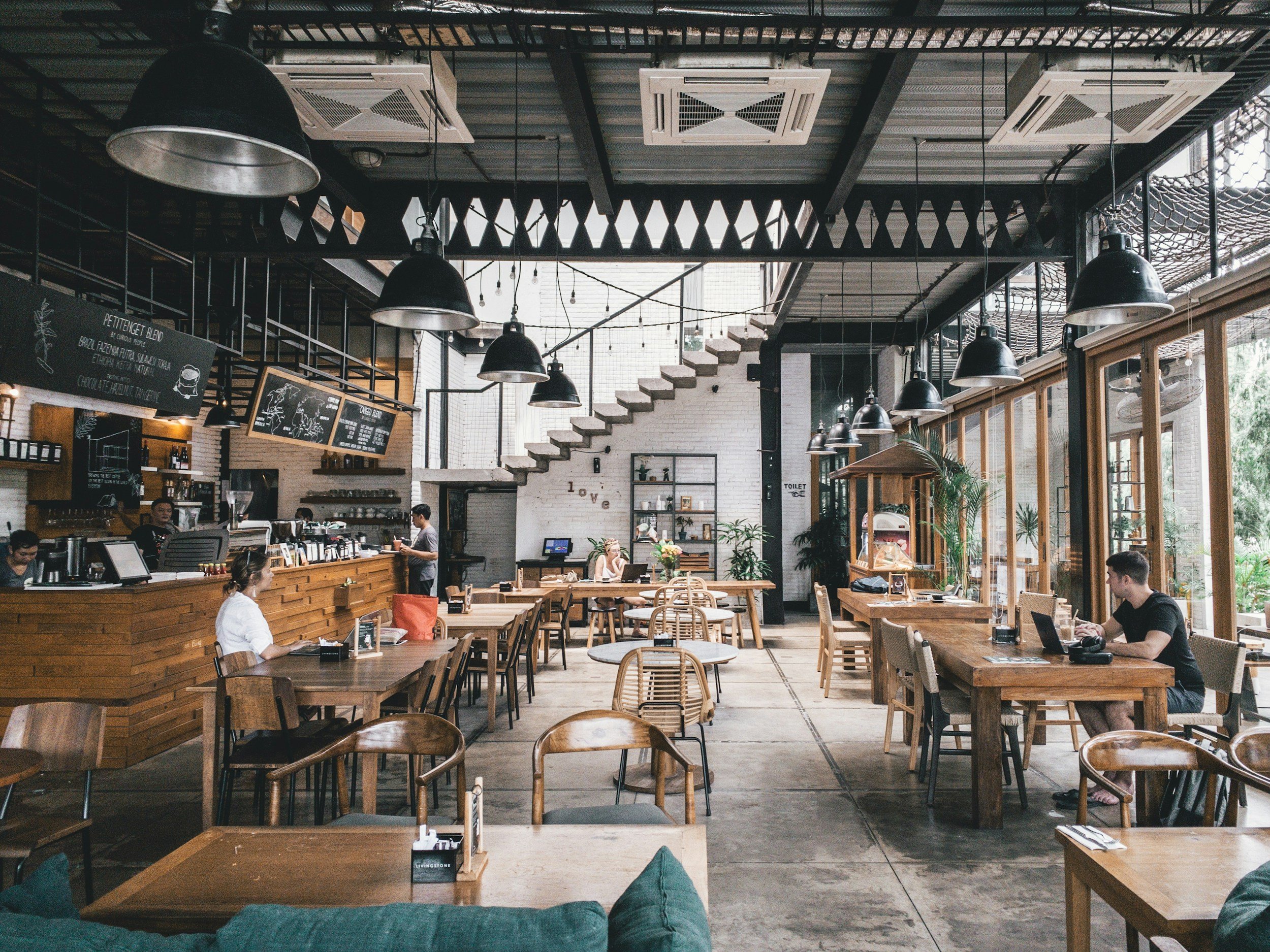 Intérieur d'un café moderne avec tables en bois, grande baie vitrée laissant entrer beaucoup de lumière naturelle, quelques clients assis travaillant ou se relaxant, présentoirs pour nourriture et boissons, et décorations vertes.