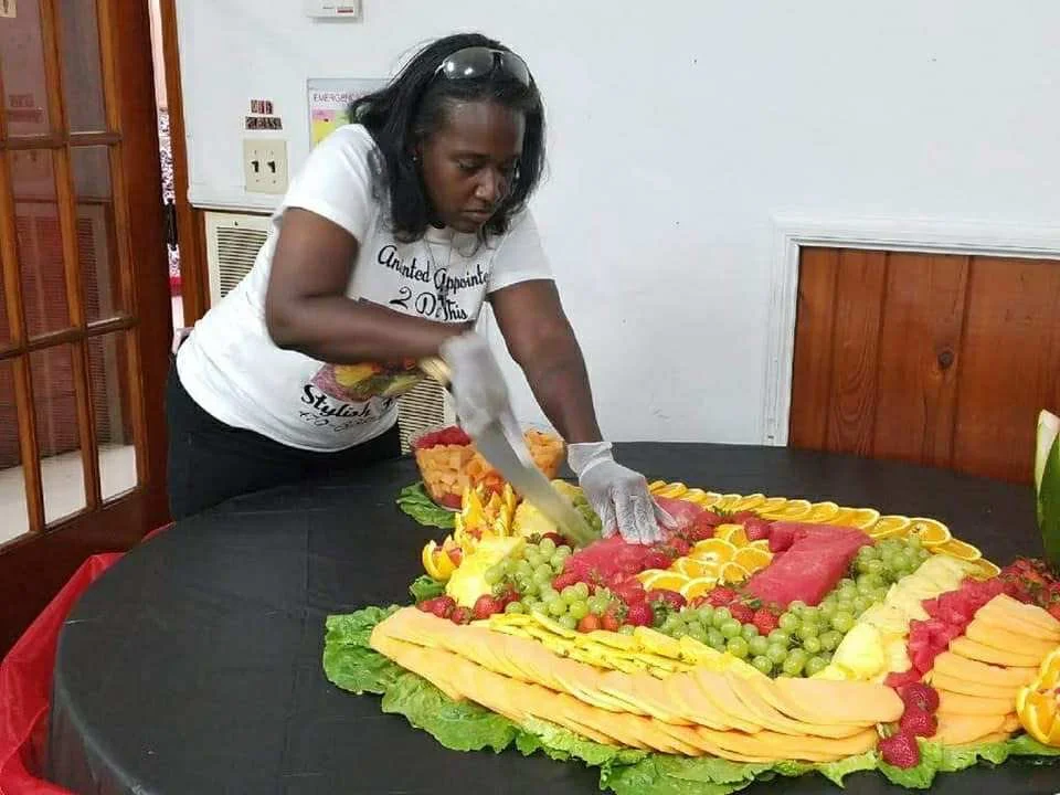 A woman is slicing a large fruit arrangement on a table, which includes slices of watermelon, cantaloupe, honeydew, and grapes, arranged on a bed of lettuce leaves.