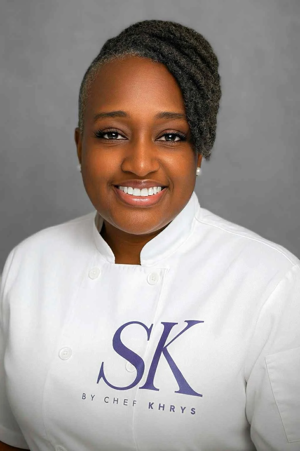 Portrait of a woman with short, styled hair, smiling, wearing a white chef's coat with the letters 'SK' and 'by Chef Khrys' printed on it, against a gray background.