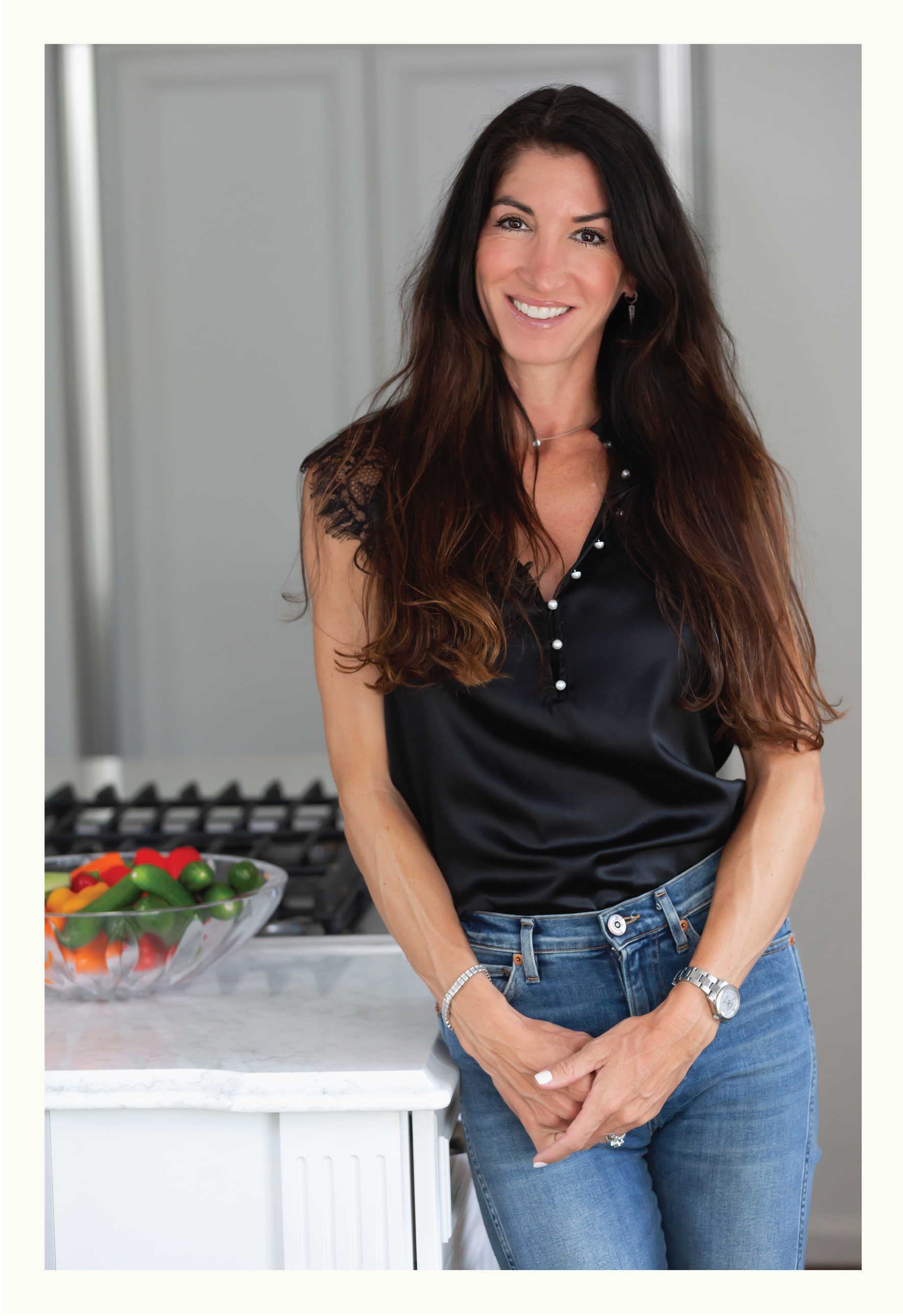 A woman with long dark hair smiling, wearing a black sleeveless top with lace details and blue jeans, standing in a kitchen next to a bowl of colorful bell peppers and cherry tomatoes.