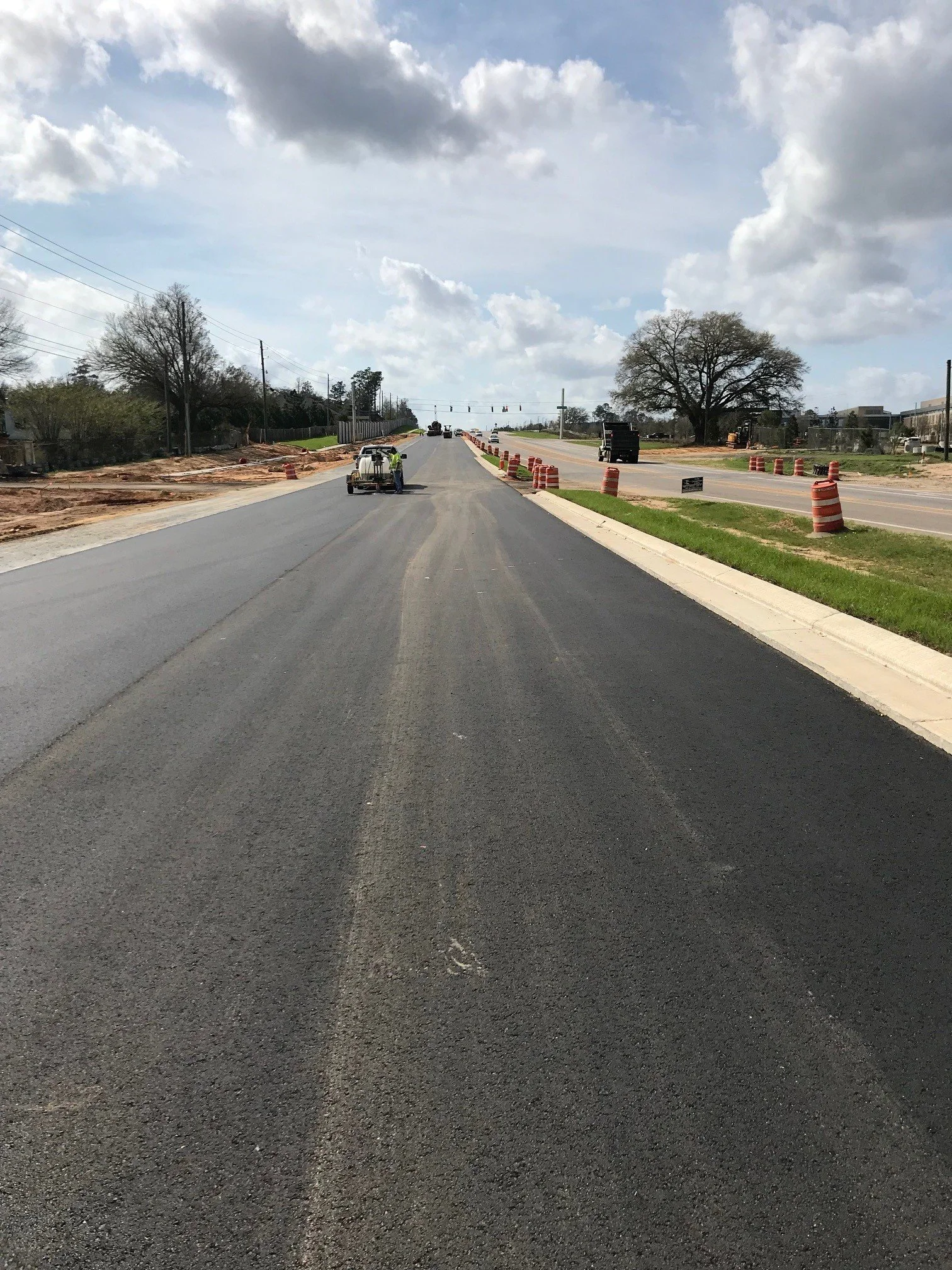 A road under construction with orange traffic barrels, a worker, and construction vehicles, with cloudy skies overhead.