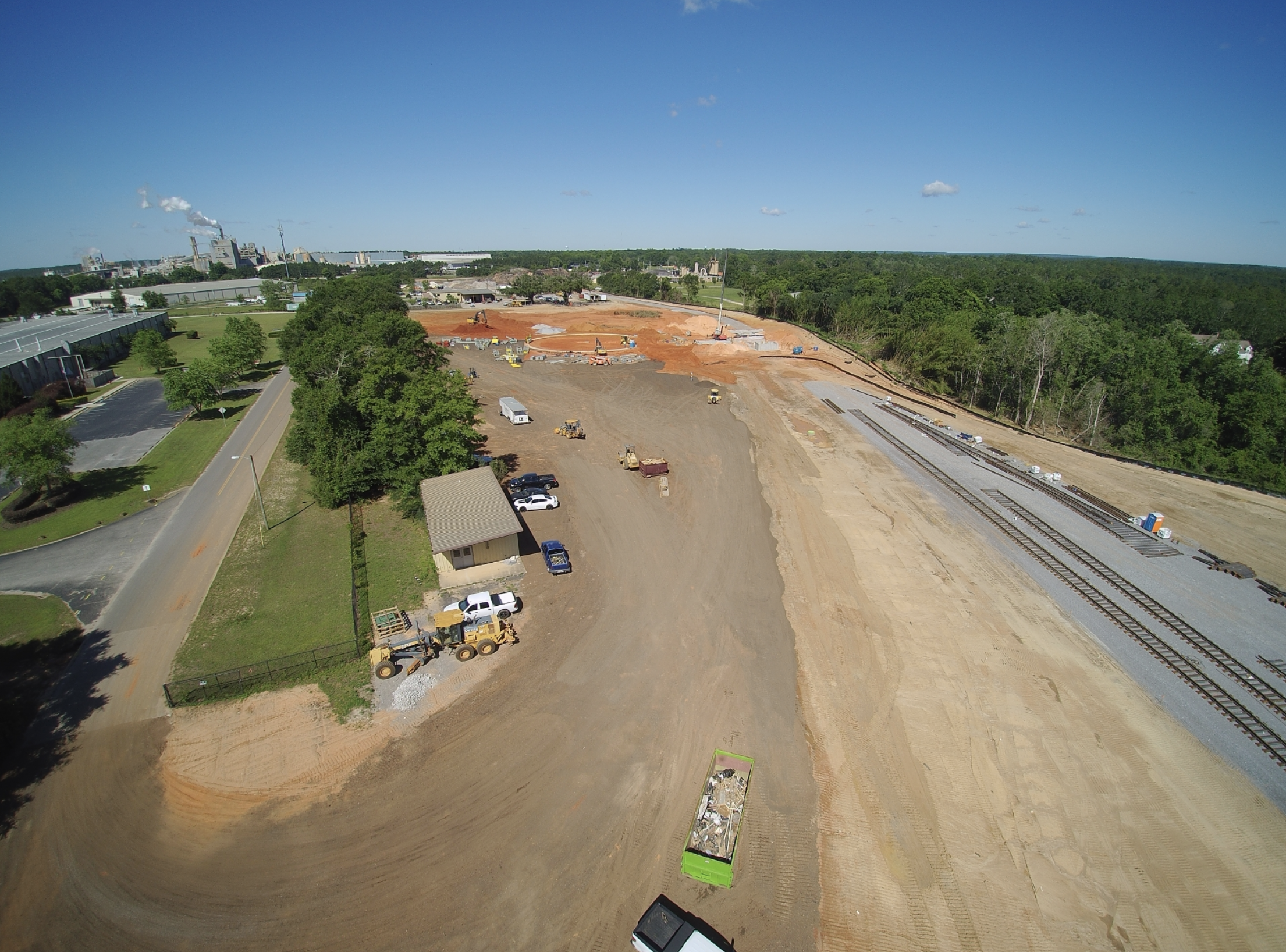 Construction site with heavy machinery and railroad tracks, green trees, and industrial buildings in the distance under a blue sky.