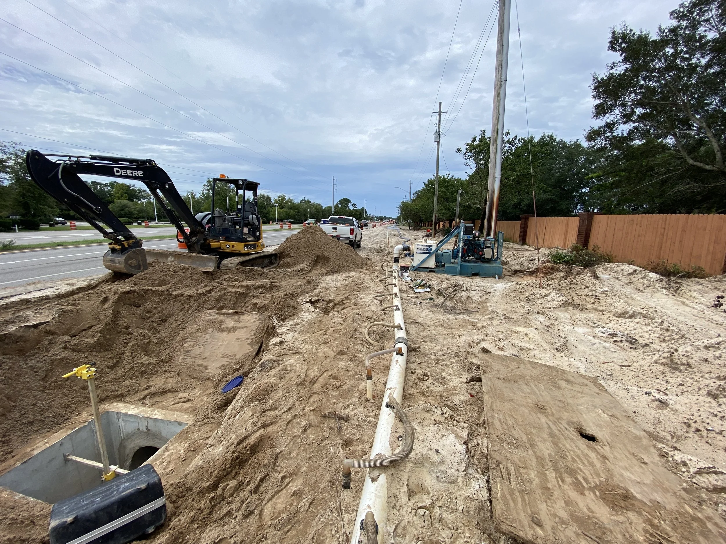 Construction site on the side of a road with a small excavator, pipes, and equipment for underground work, and a wooden fence in the background.