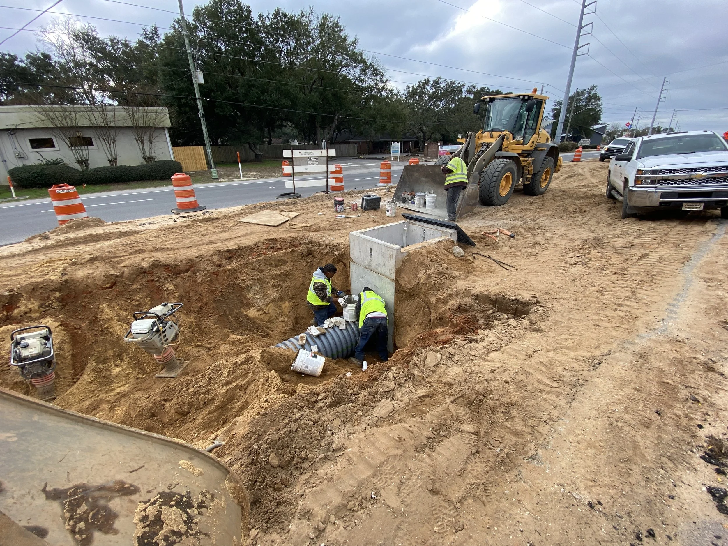 Construction workers installing underground utility piping next to a road, with construction equipment, vehicles, and traffic cones present.