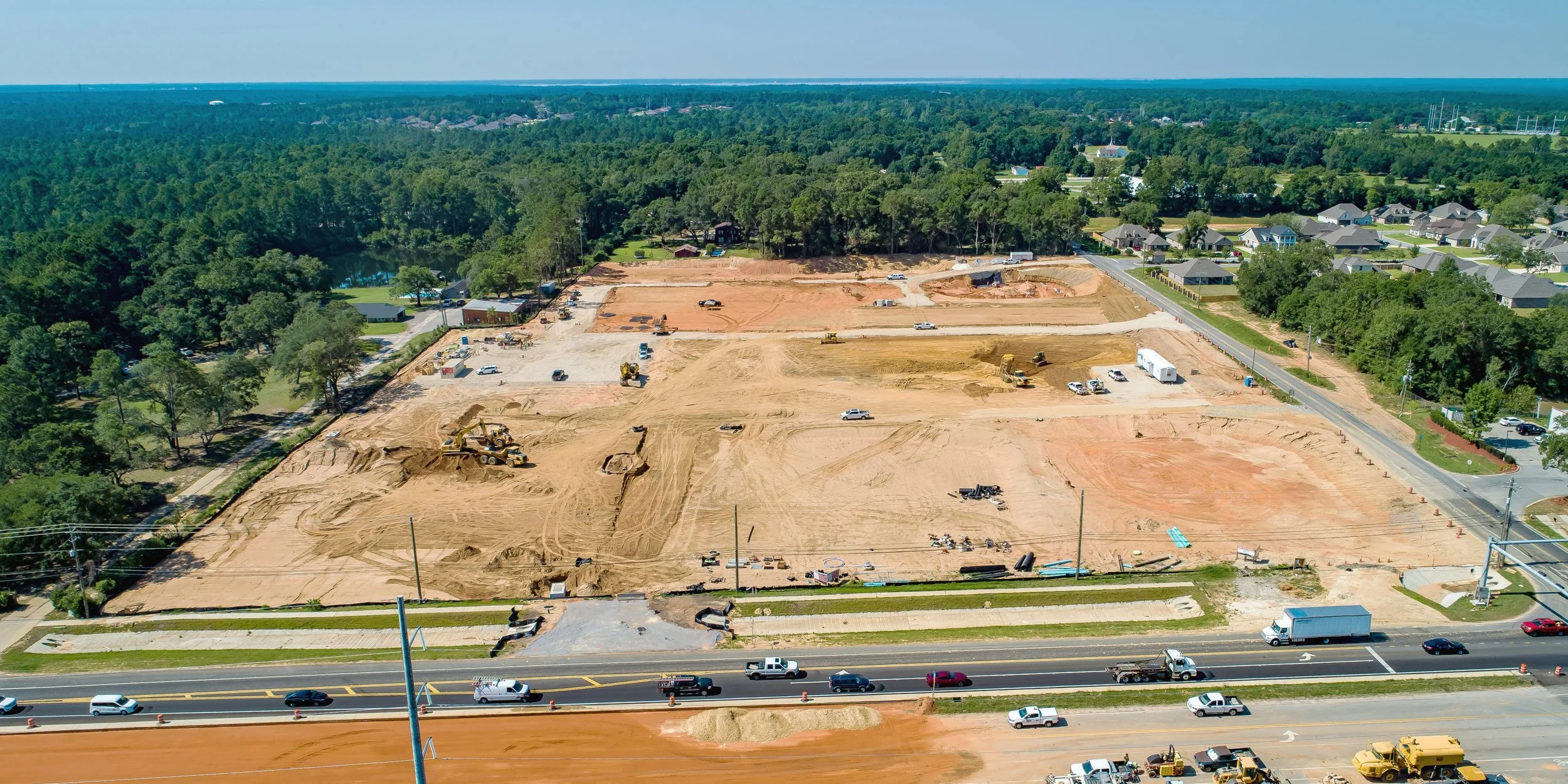 Aerial view of a construction site with machinery and vehicles, adjacent to a residential neighborhood and a forested area.