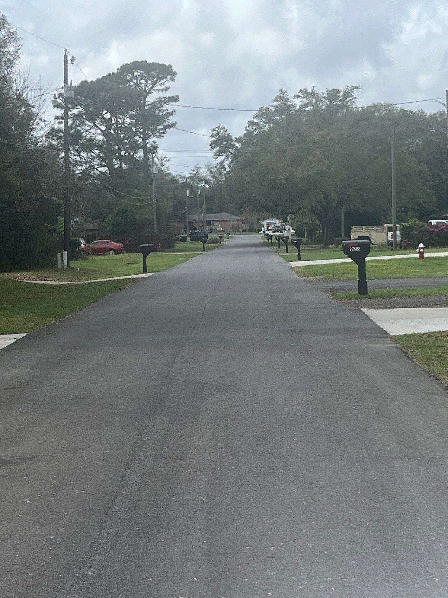 A suburban residential street with lined up mailboxes along the pavement and houses with trees in the background, under cloudy skies.