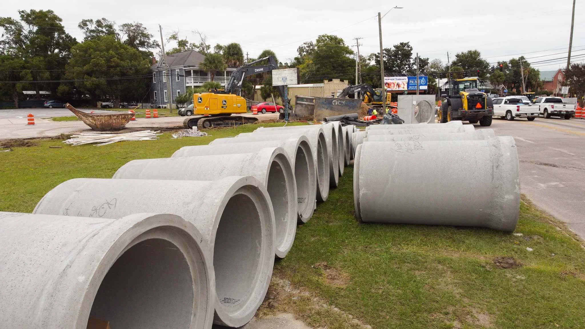 Construction site with large concrete pipes lined up on the grass, construction equipment, and vehicles on the street.