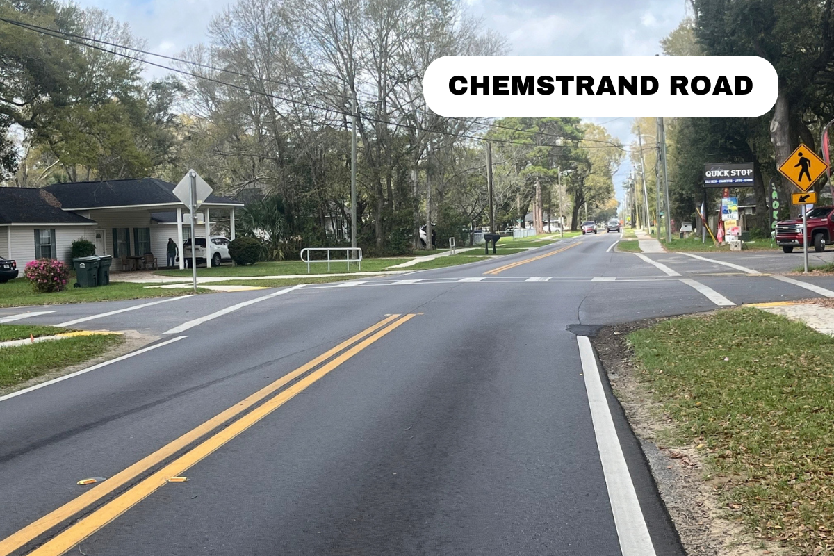 A street view of Chemstrand Road with a pedestrian crosswalk, traffic signs, and trees lining the sides of the road.