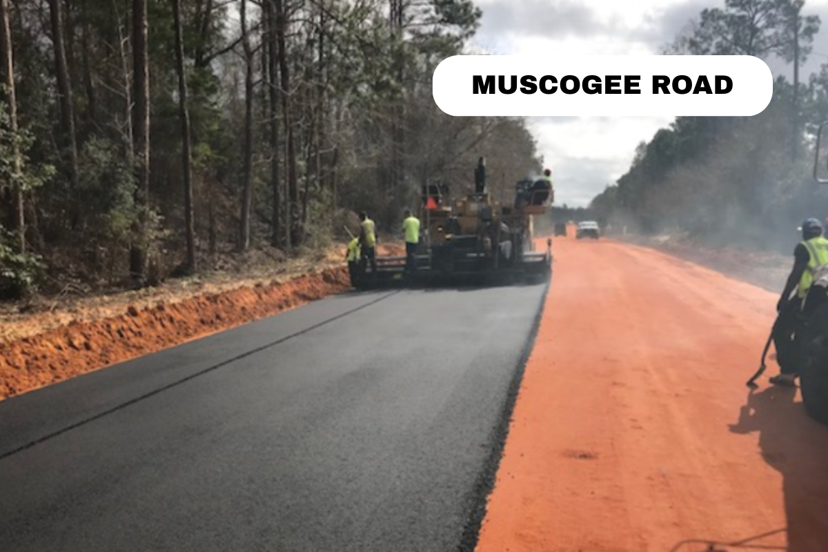 Workers paving a road on Muscogee Road with asphalt, with trees on the side and a vehicle in the background.