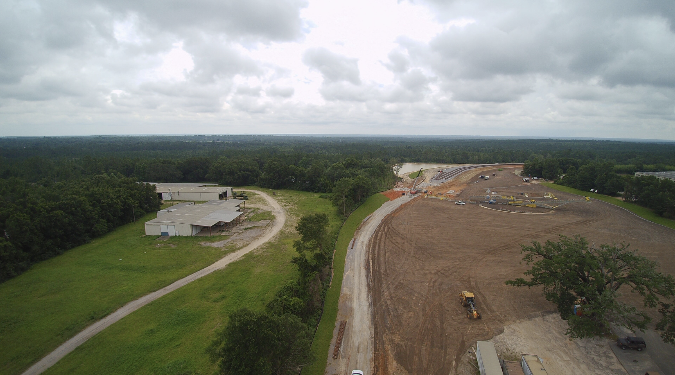 Construction site alongside a green, wooded area, with ongoing earthwork, construction vehicles, and partially built infrastructure under a cloudy sky.