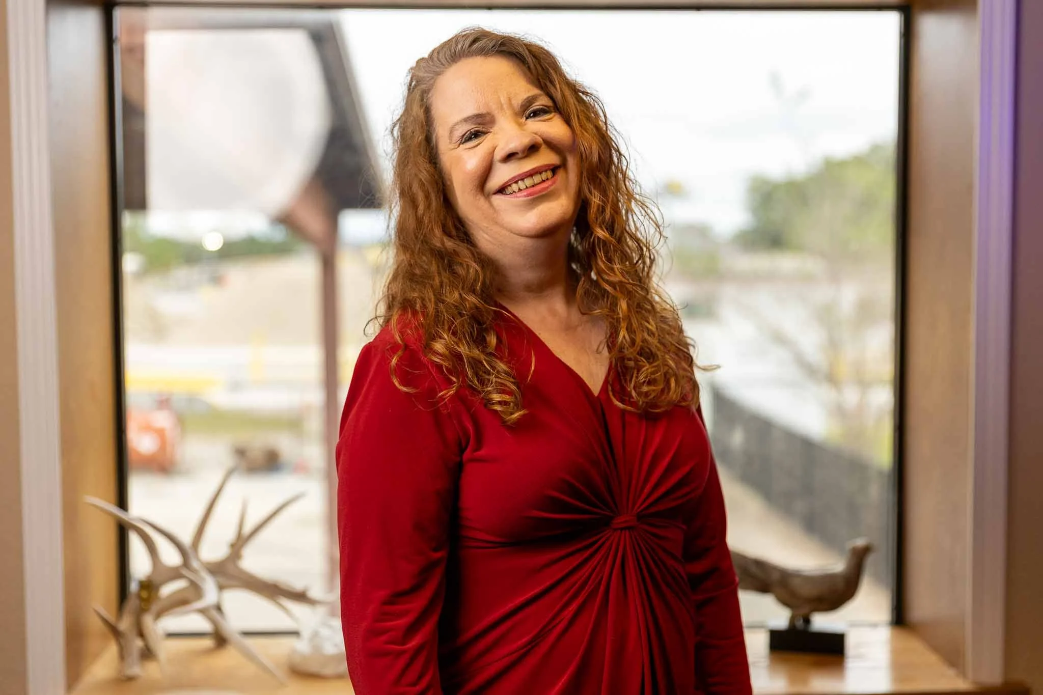 A woman with curly red hair wearing a red dress smiling while standing indoors in front of a window with outside view.