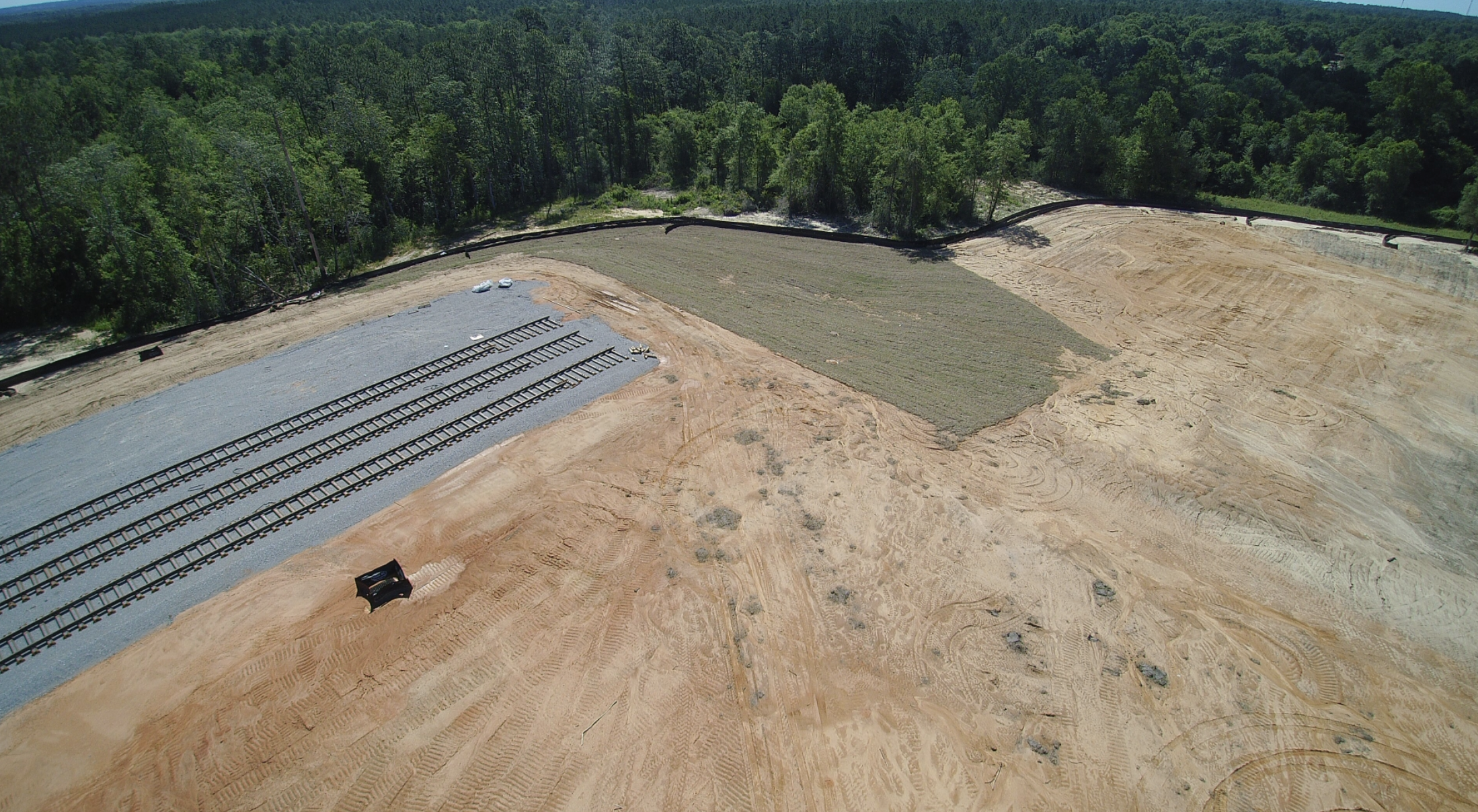 An aerial view of a construction site with train tracks being built, cleared earth, and nearby forested area.