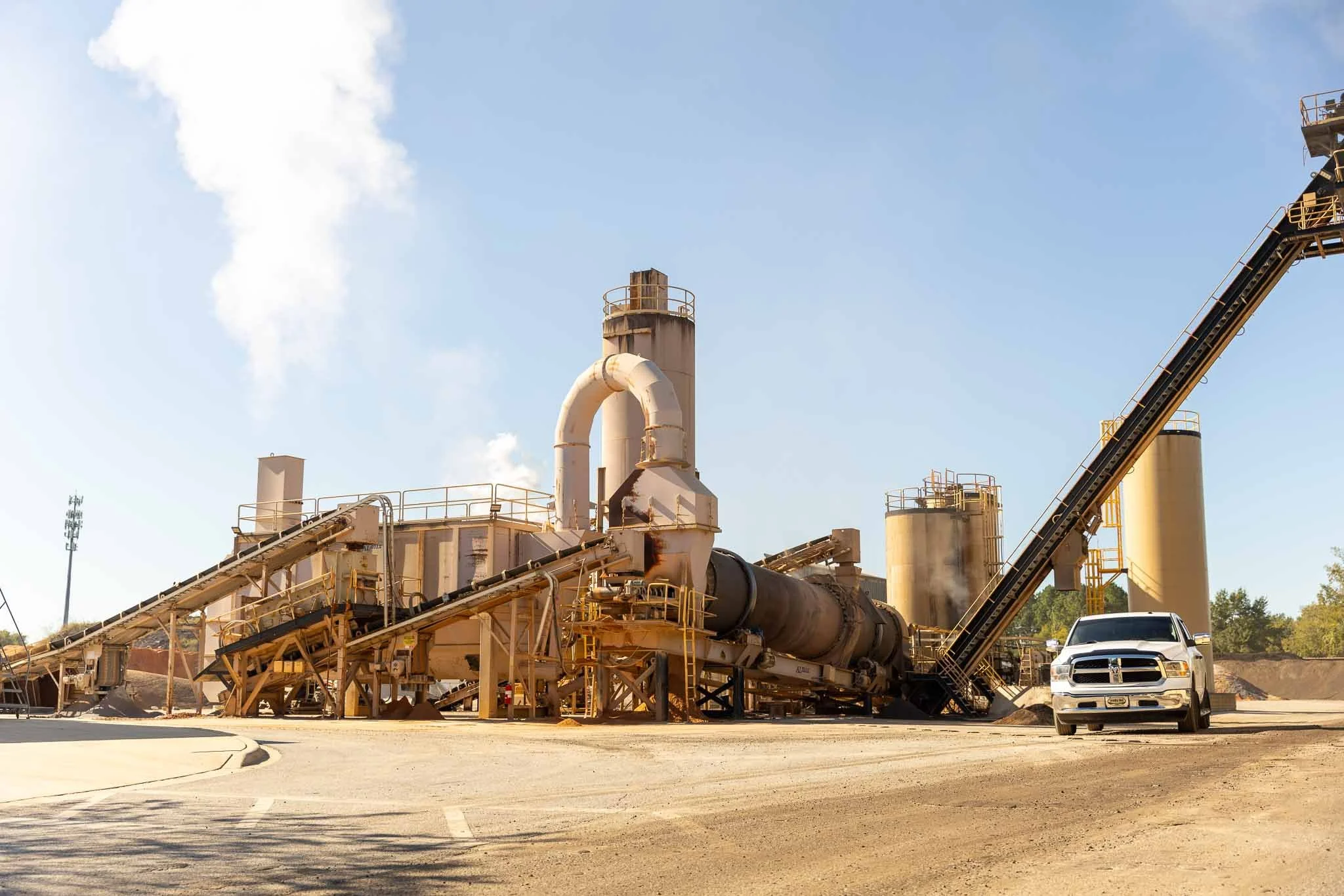 View of an industrial plant with smokestacks, conveyor belts, and a white pickup truck parked nearby.