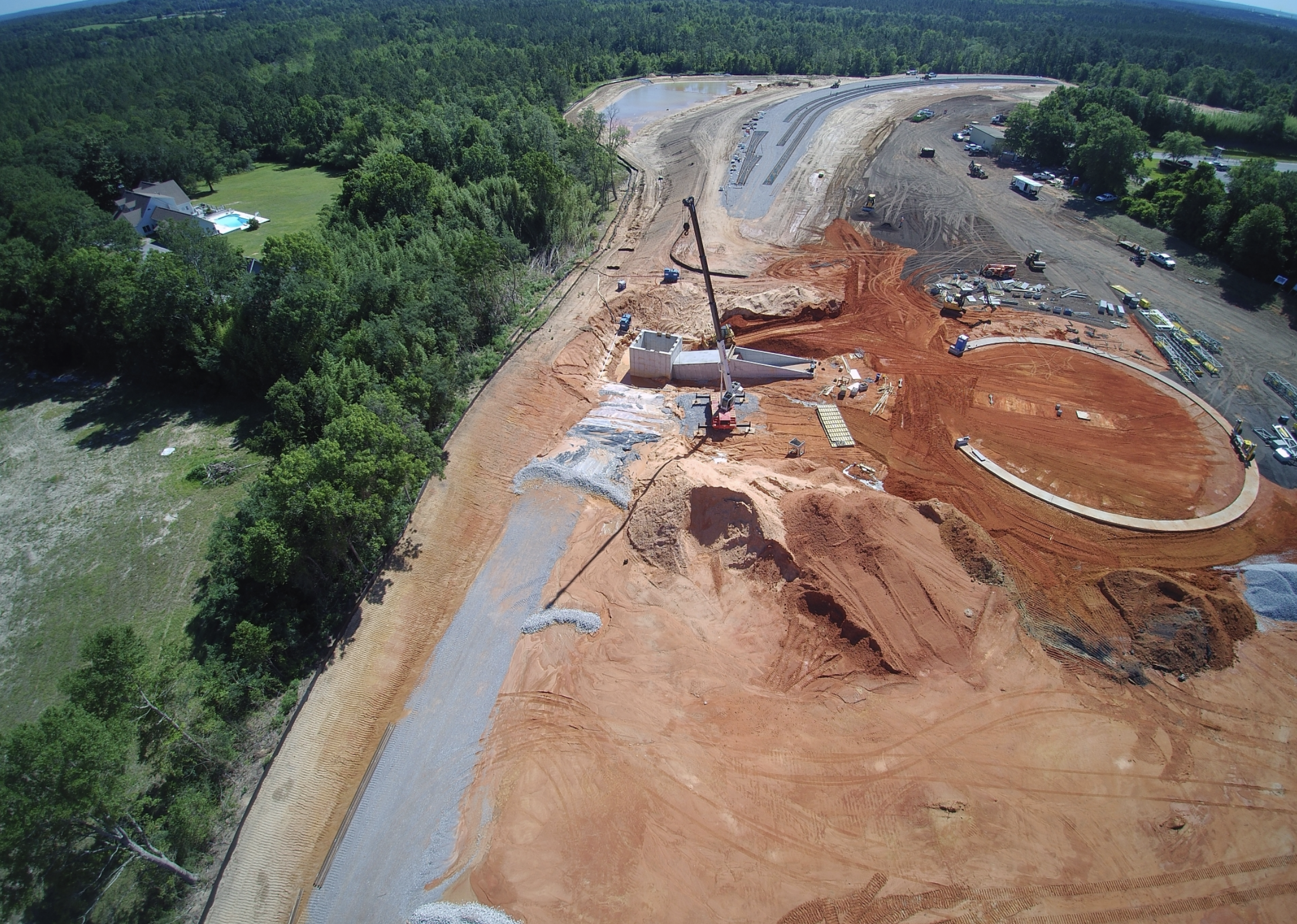 Aerial view of a construction site with dirt and gravel pathways, a crane, and circular structure being built, surrounded by trees and some nearby houses.