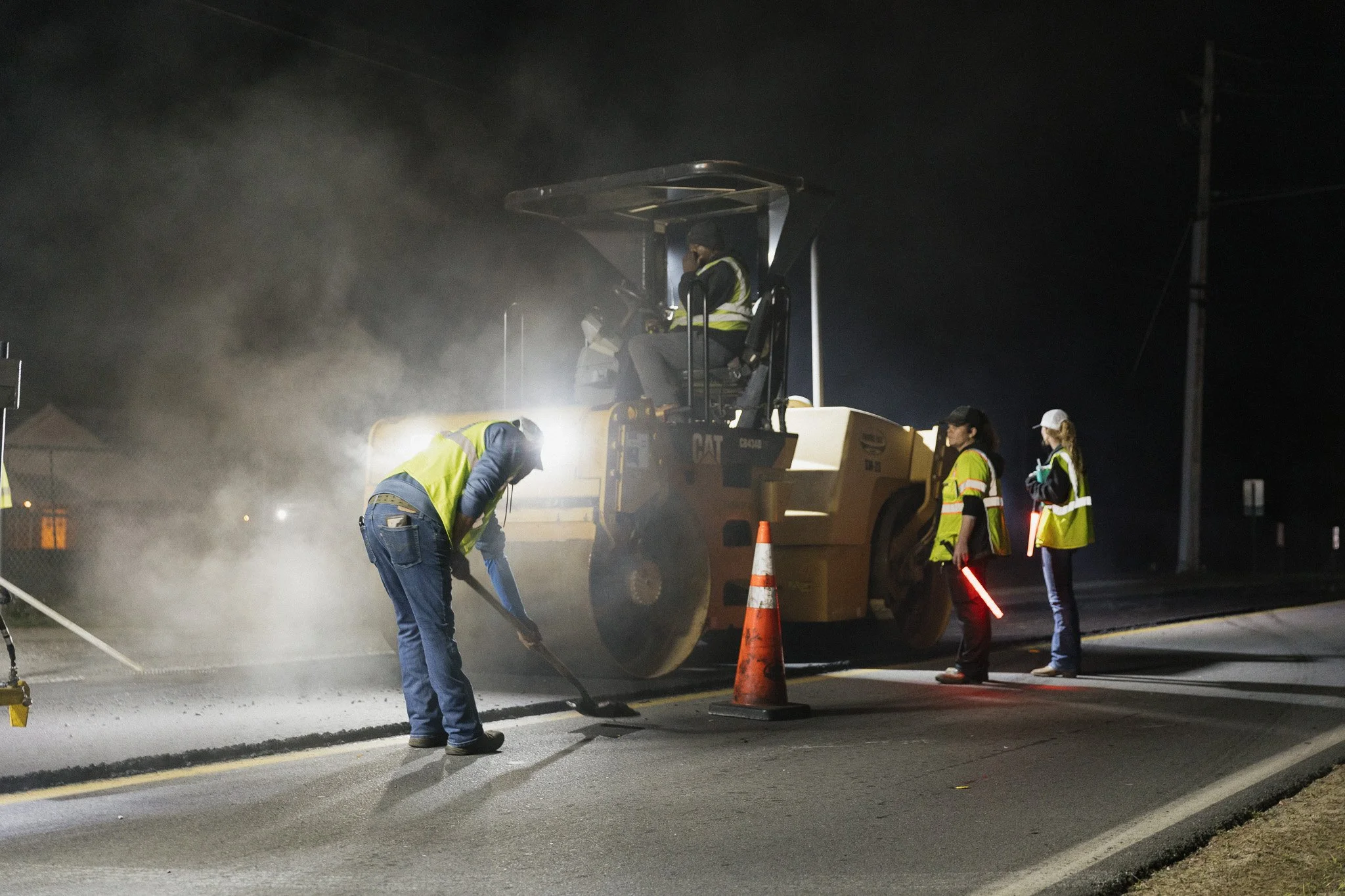A large yellow road roller compacts asphalt on a road construction site, with trees and utility poles in the background.