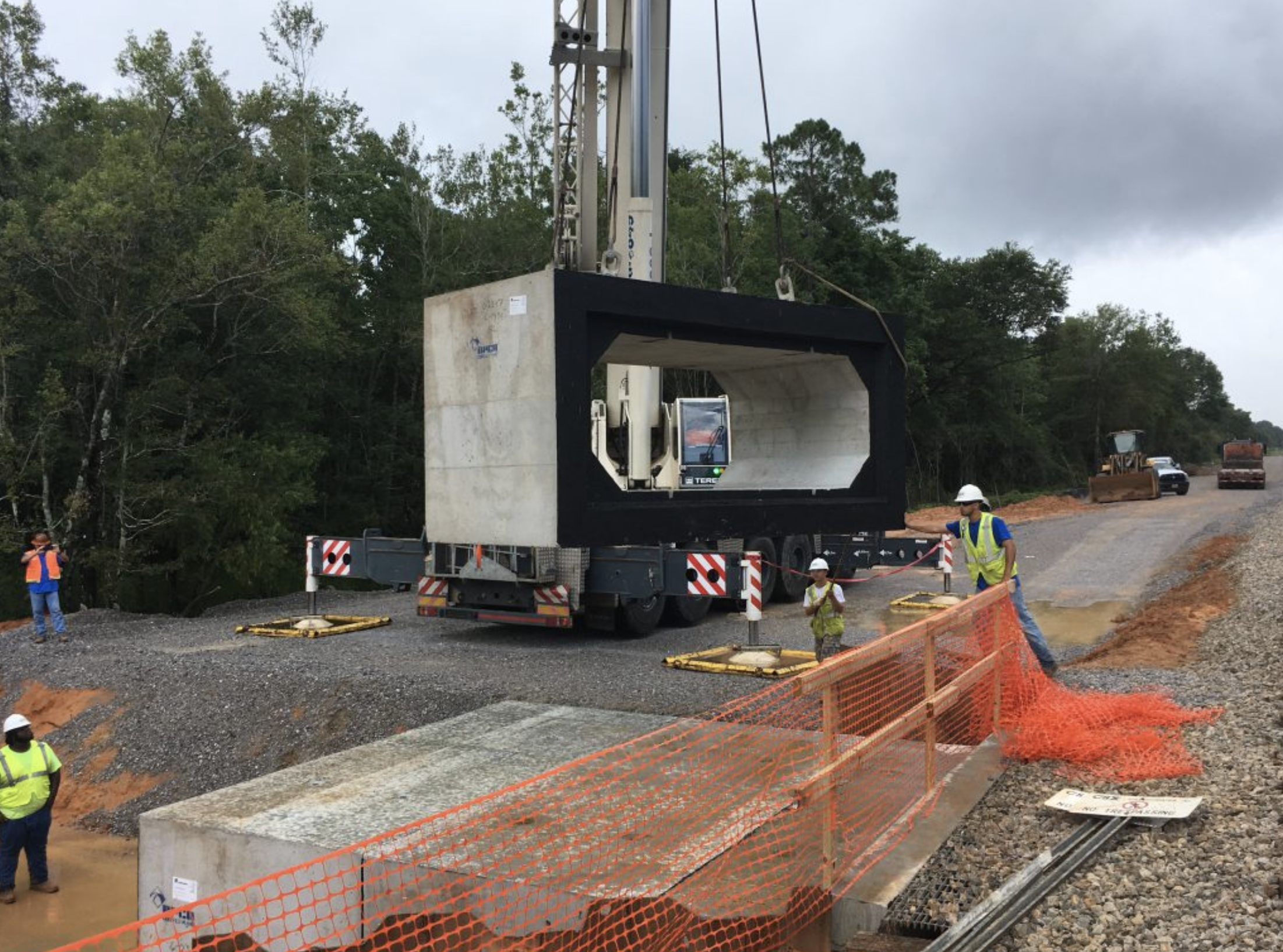 Construction workers and heavy machinery working on a road project in a rural area, with trees in the background and construction equipment in use.
