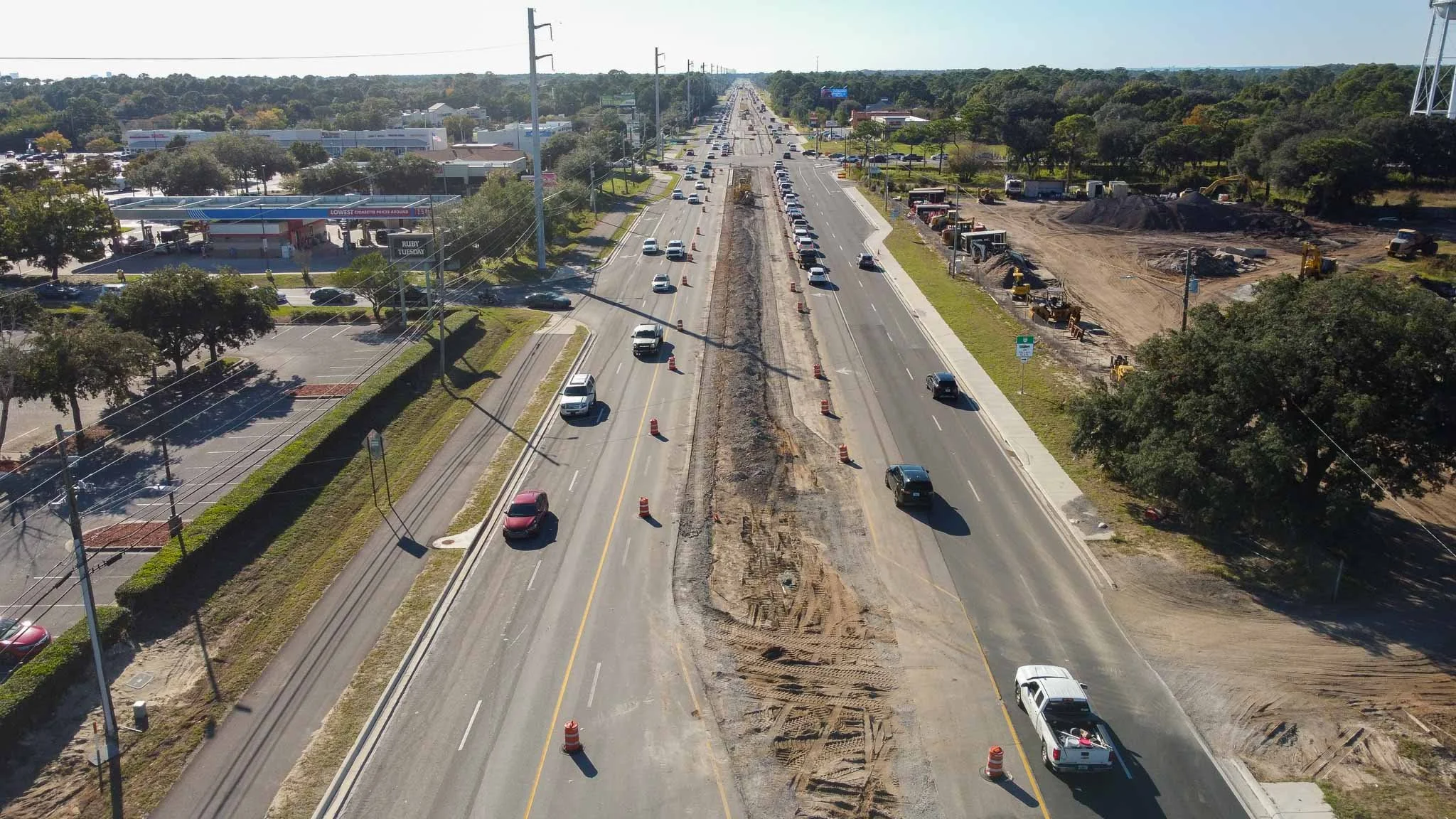 Aerial view of a multi-lane road under construction, with orange traffic cones and construction equipment on the right side, and traffic moving in both directions.