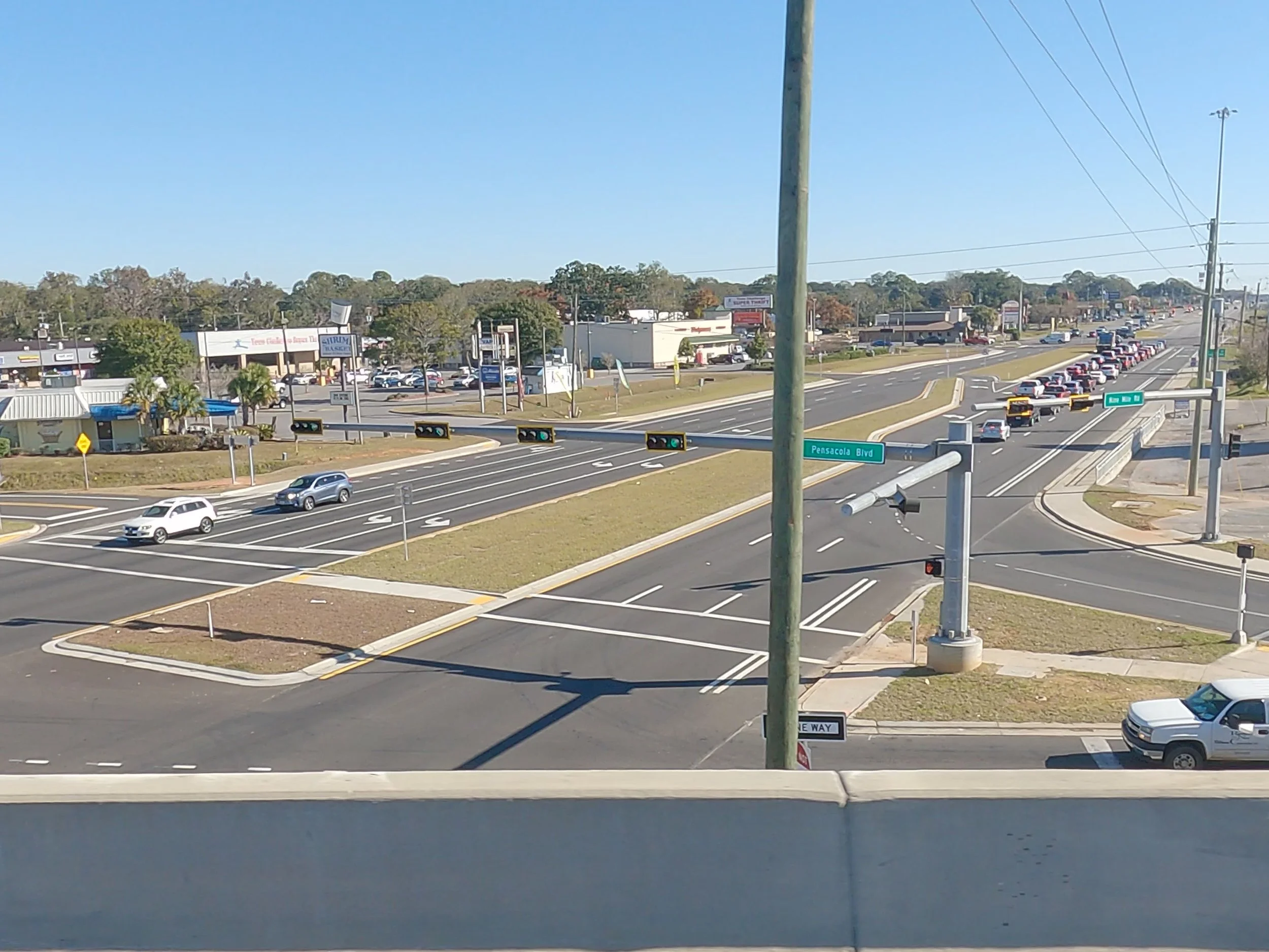 Intersection view of Pensacola Blvd with cars lined up at traffic lights and a line of cars waiting to turn. Street signs indicate Pensacola Blvd and one-way street. Various commercial buildings and parking lots are visible in the background, along w