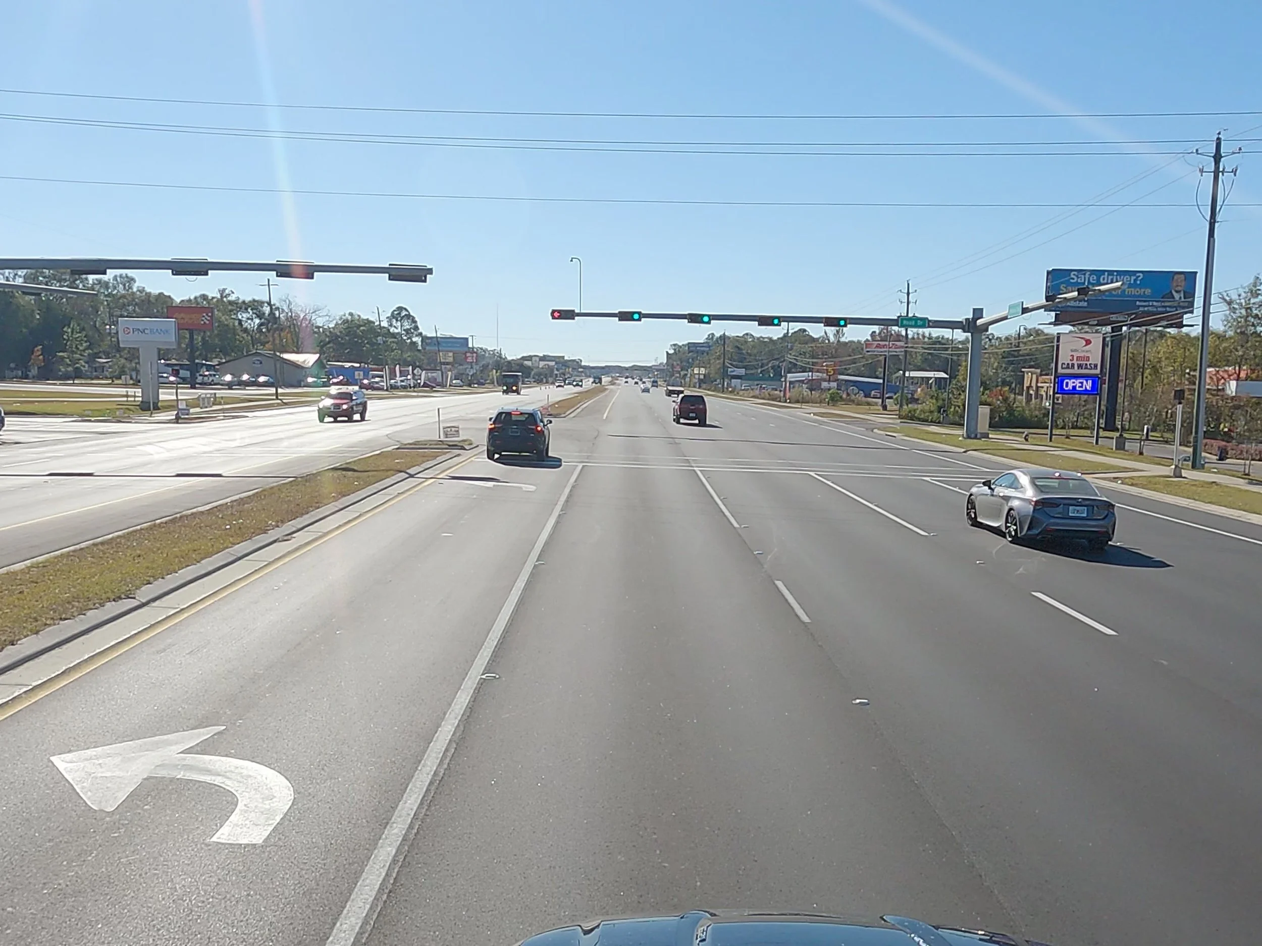 A wide view of a multi-lane road with cars, traffic lights, billboards, and a clear blue sky. A car is making a left turn marked by a white left turn arrow on the road.